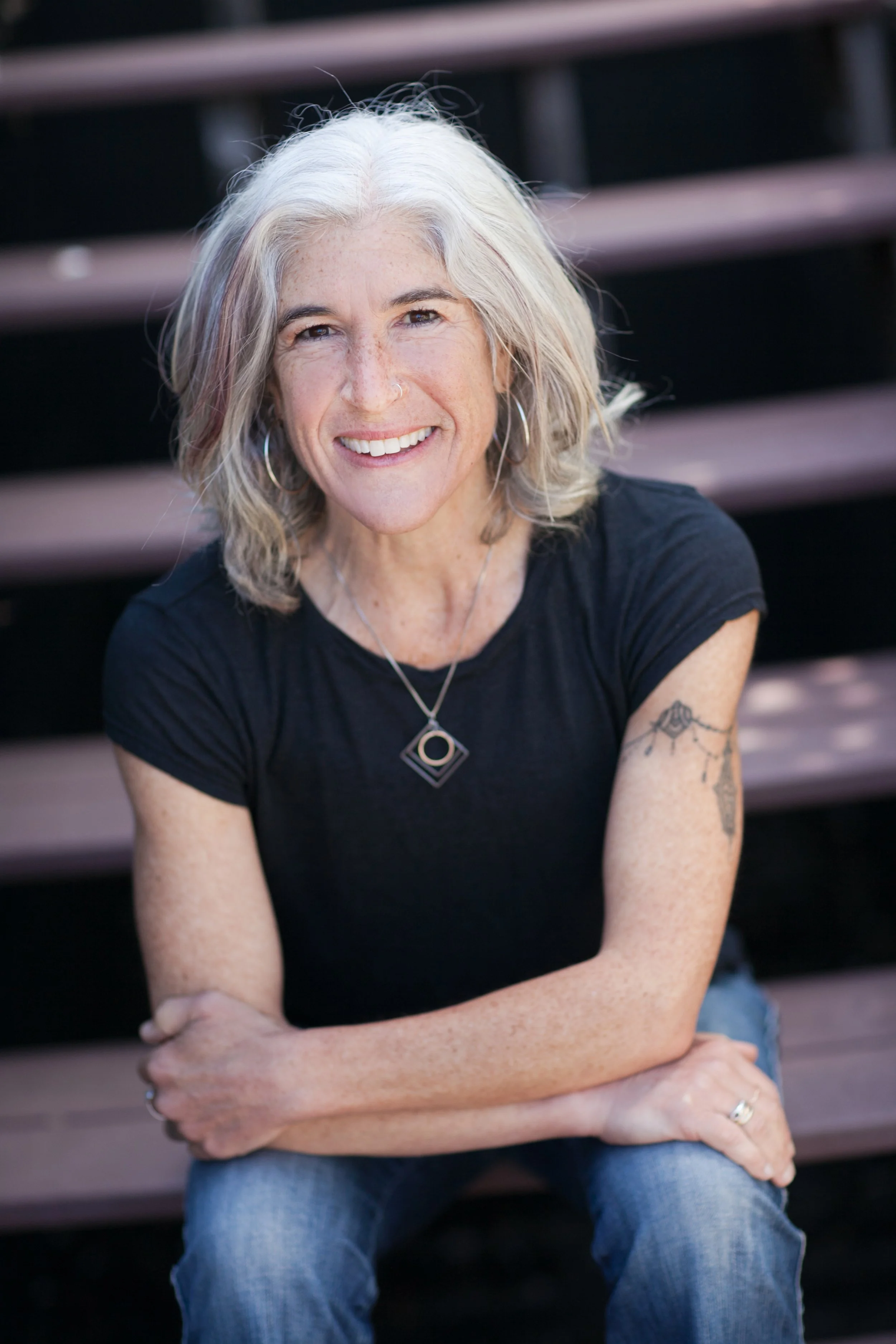A photo of Rachel Heron; a woman smiling and sitting outdoors on wooden steps. She has grey hair, a nose ring, visible freckles, and a tattoo on her left arm.
