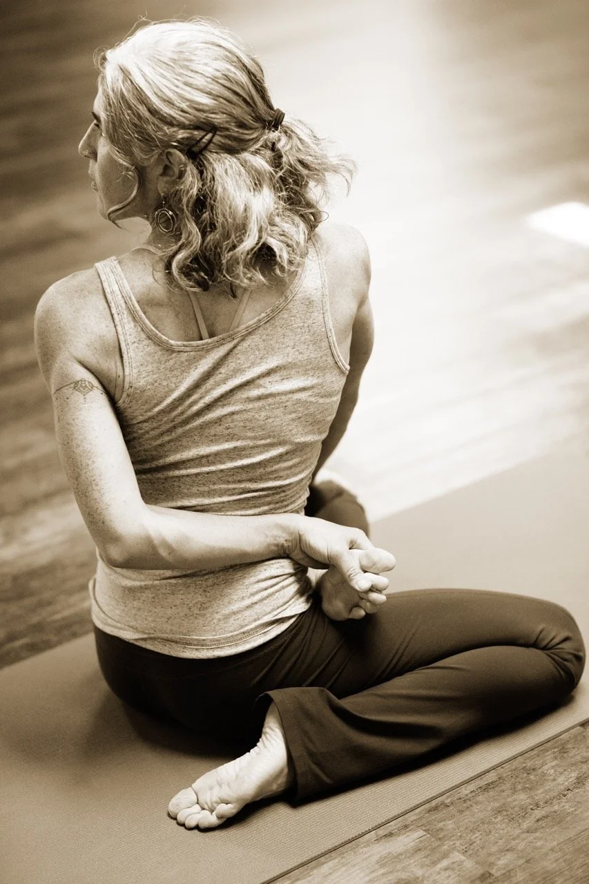 A photo of Rachel Heron practicing yoga, in a seated twist pose, cross-legged on a mat with her hands clasped behind her back, wearing a sleeveless top and pants.