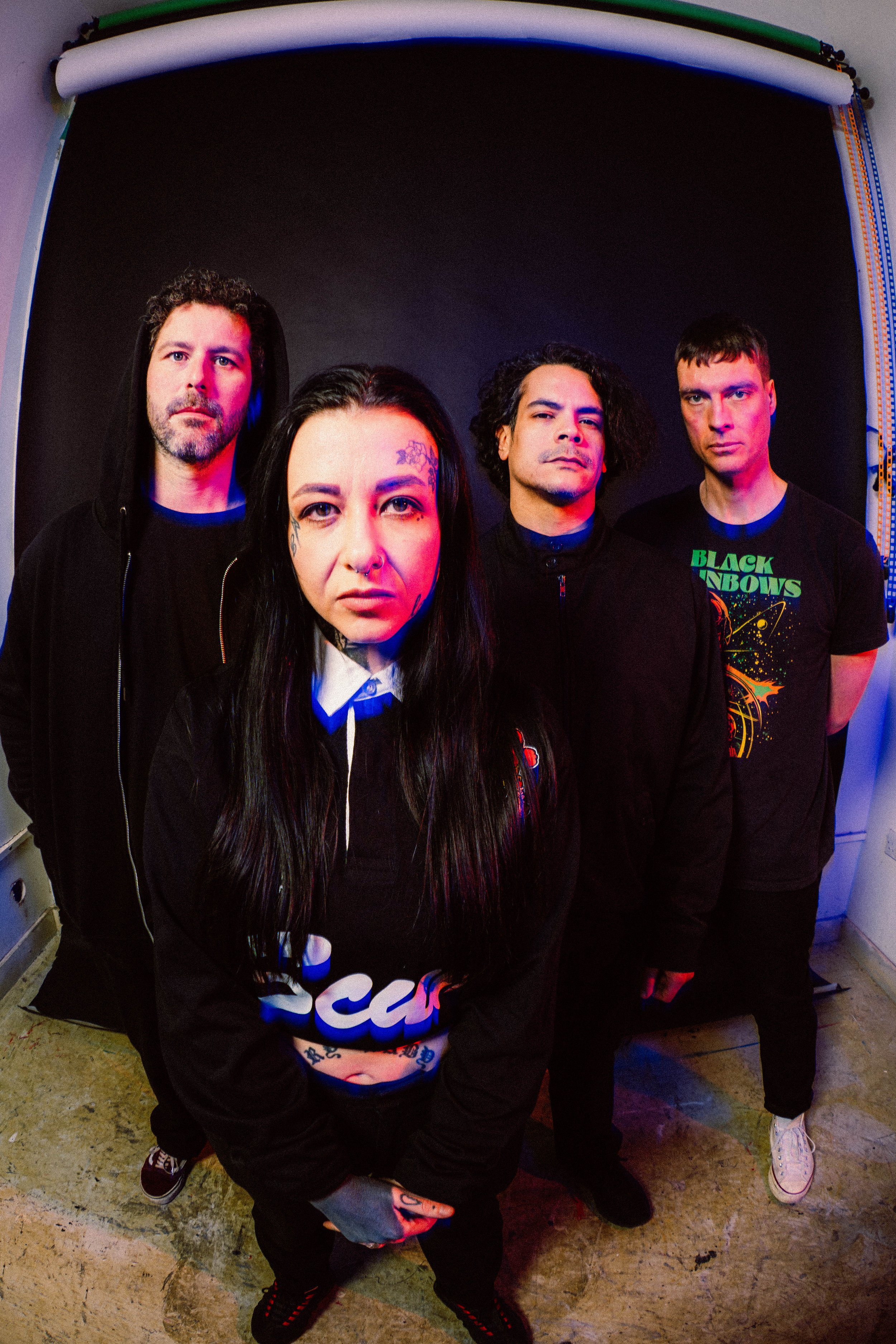 Four band members posing in front of a black backdrop, with neon lighting highlighting their faces, including female with tattoos and long dark hair, and three males with varying hairstyles and casual clothing.
