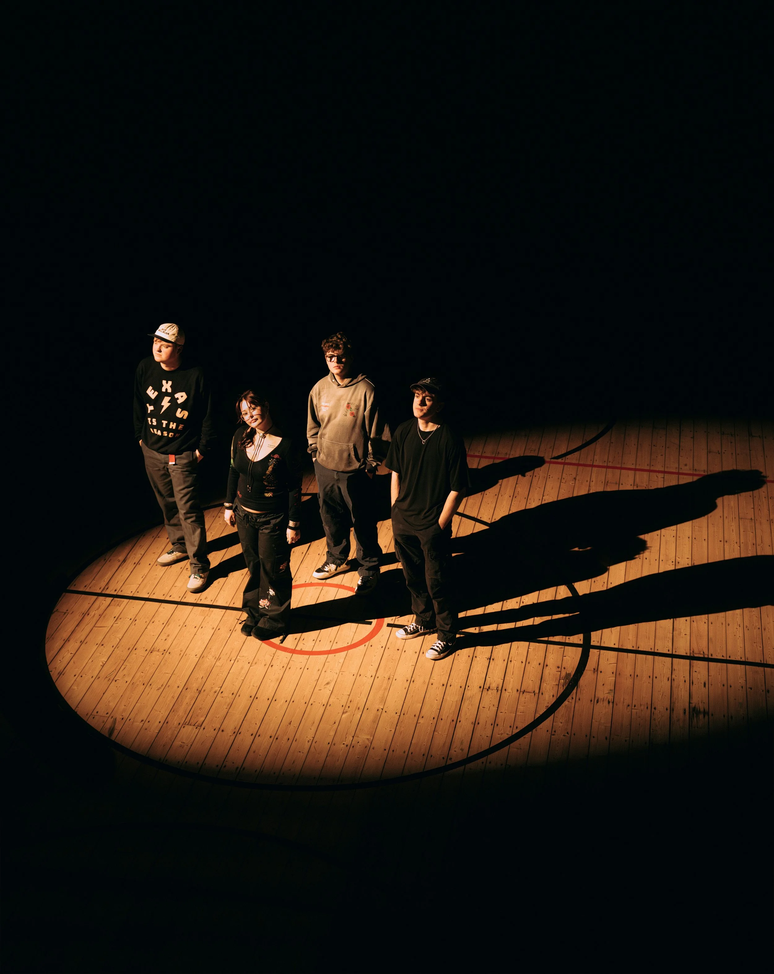 Four people standing on a basketball court with dramatic spotlight and shadows, no audience visible