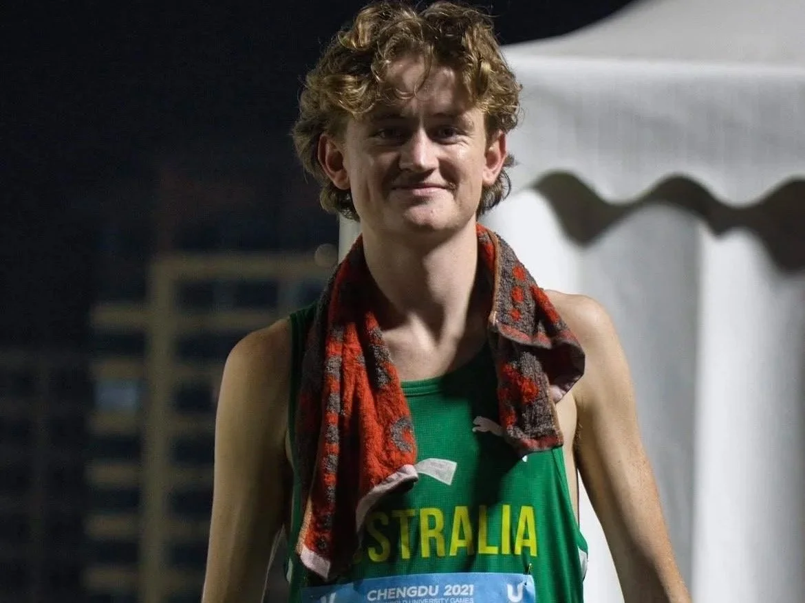 Young male athlete with curly blonde hair, wearing a green Australia sports jersey and a red patterned towel draped around his neck, smiling after a race or event at night.