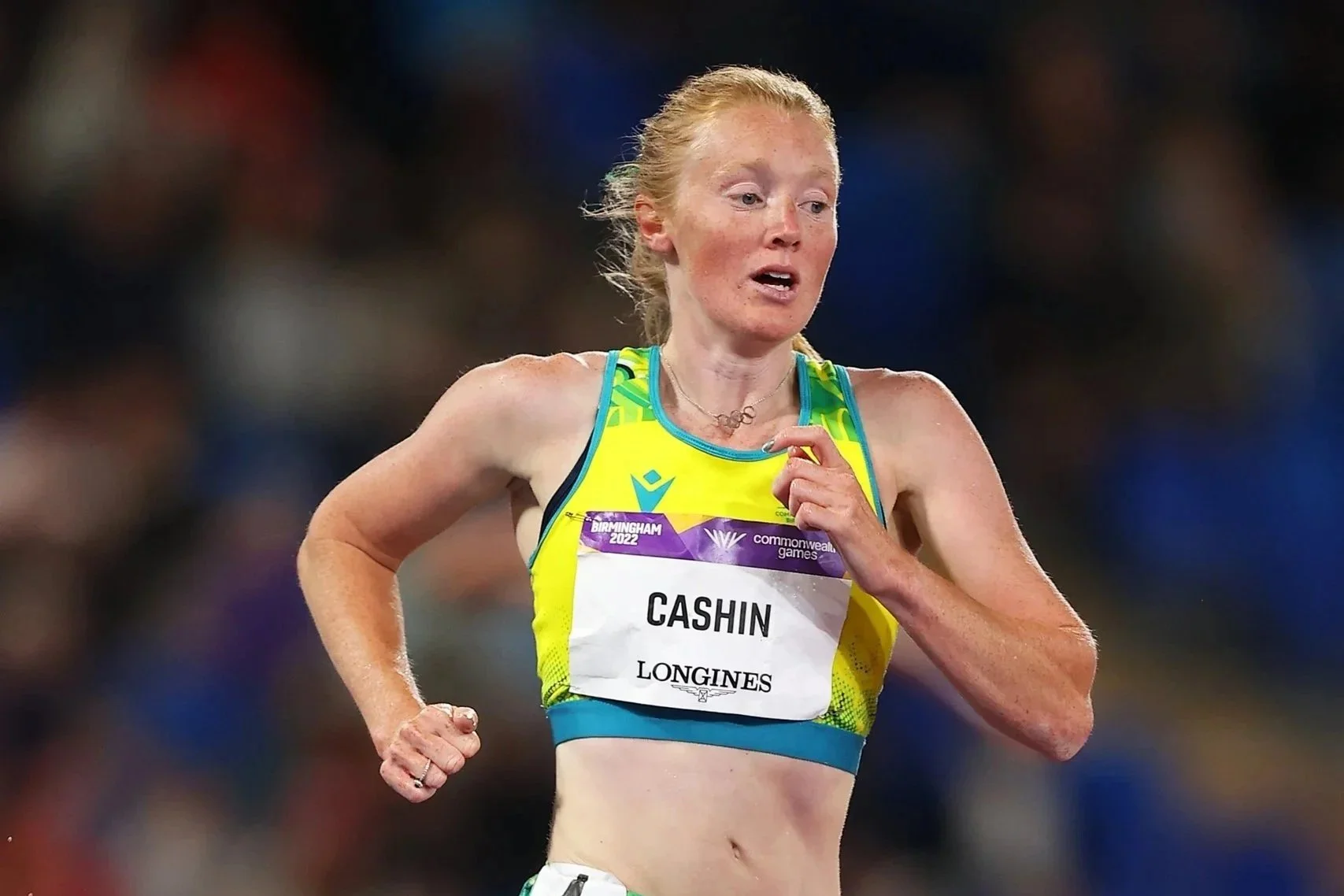 A female athlete running in a race, wearing a yellow and blue sports bra with a bib labeled 'CASHIN' and 'LONGINES', at the Birmingham 2022 Commonwealth Games.