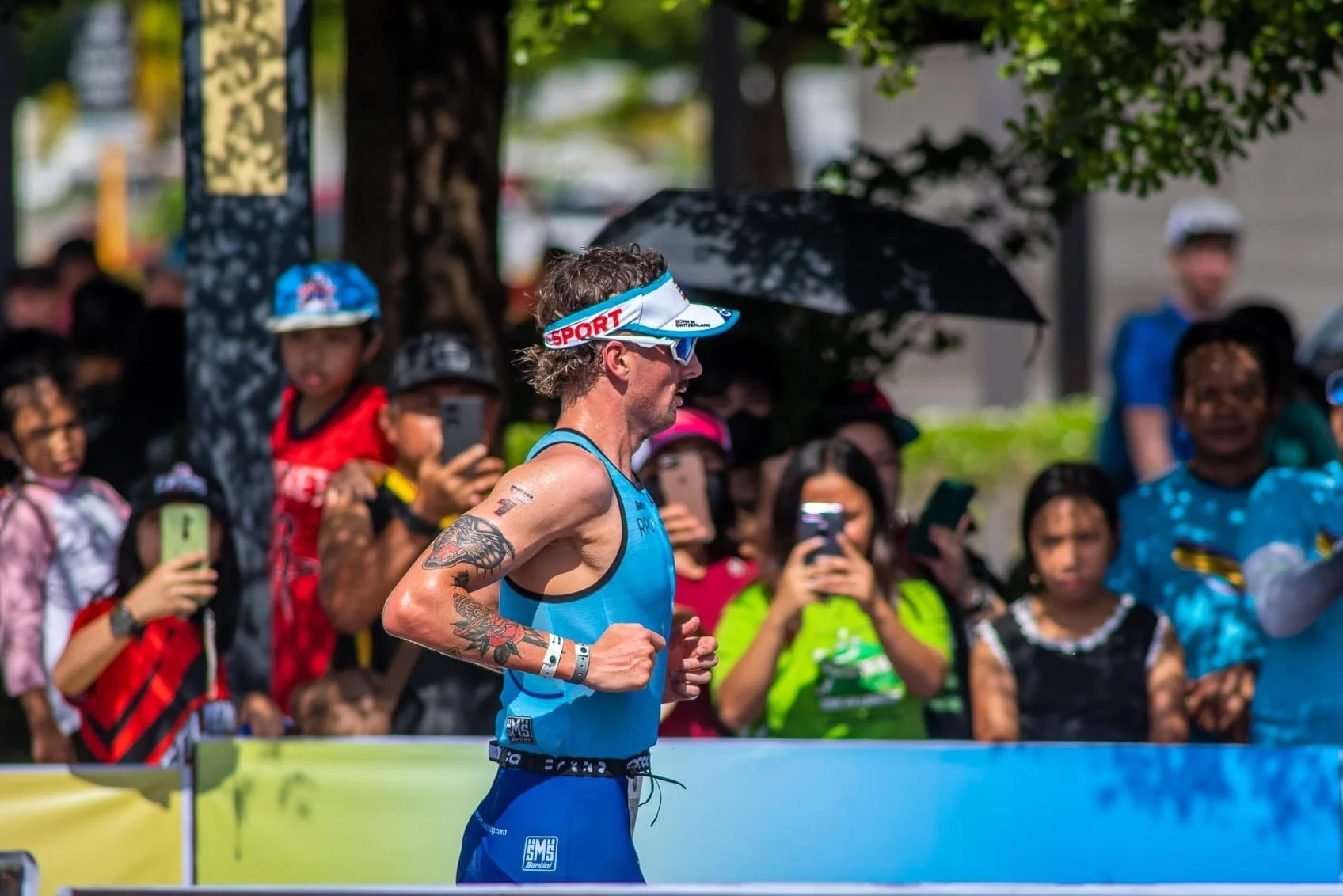 A male athlete running in a race with a crowd of spectators taking photos and watching along the side of the race course. The runner is wearing a blue athletic outfit, a visor, and sunglasses.