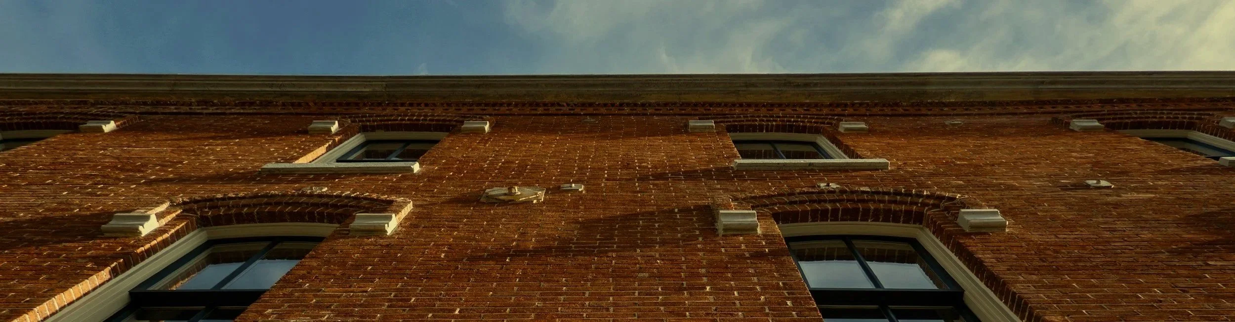 Looking up at a brick building with multiple windows and white trim, against a partly cloudy sky.