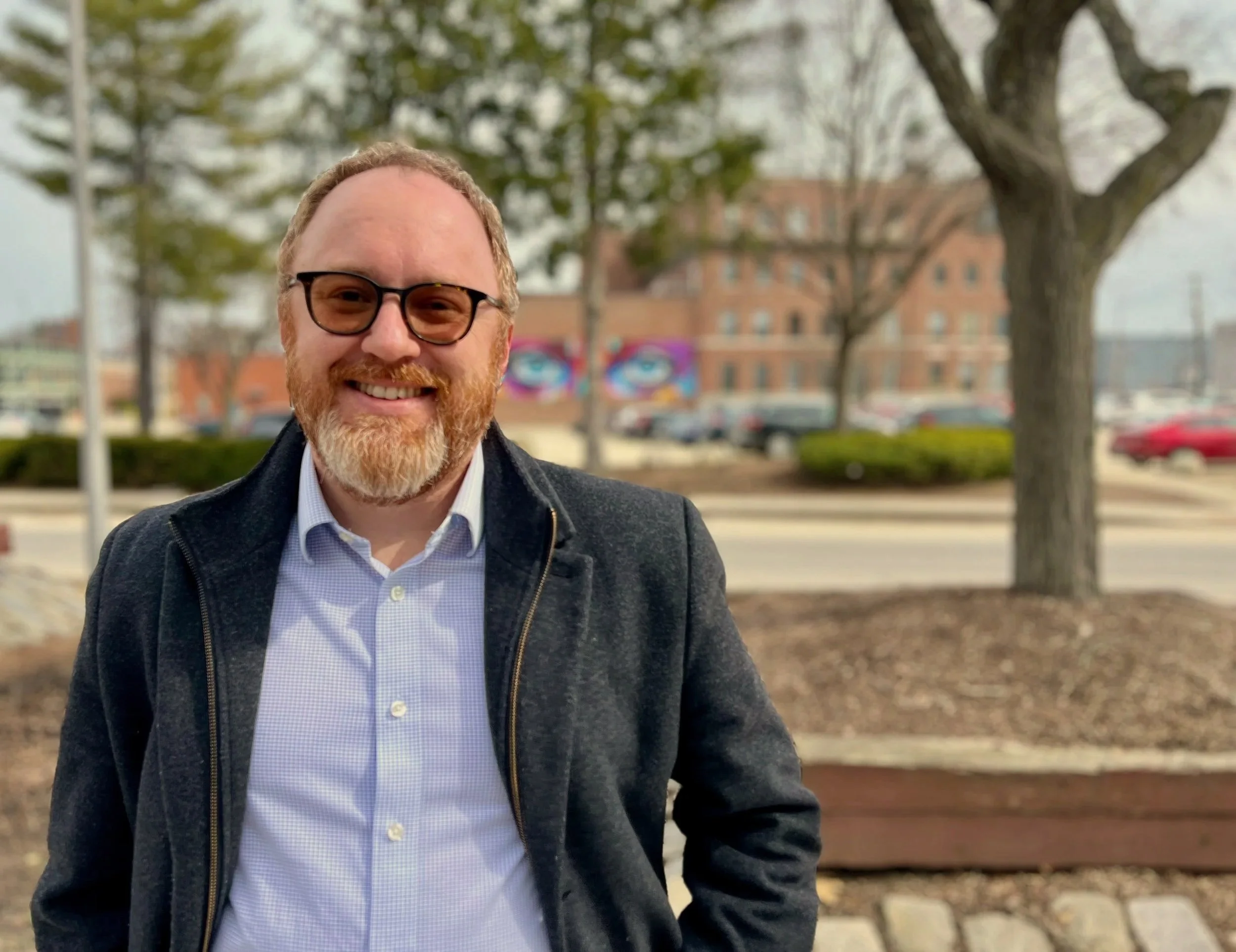 A smiling man with a beard and glasses, wearing a dark jacket and light blue shirt, standing outdoors in front of trees, a building, and parked cars on a cloudy day.