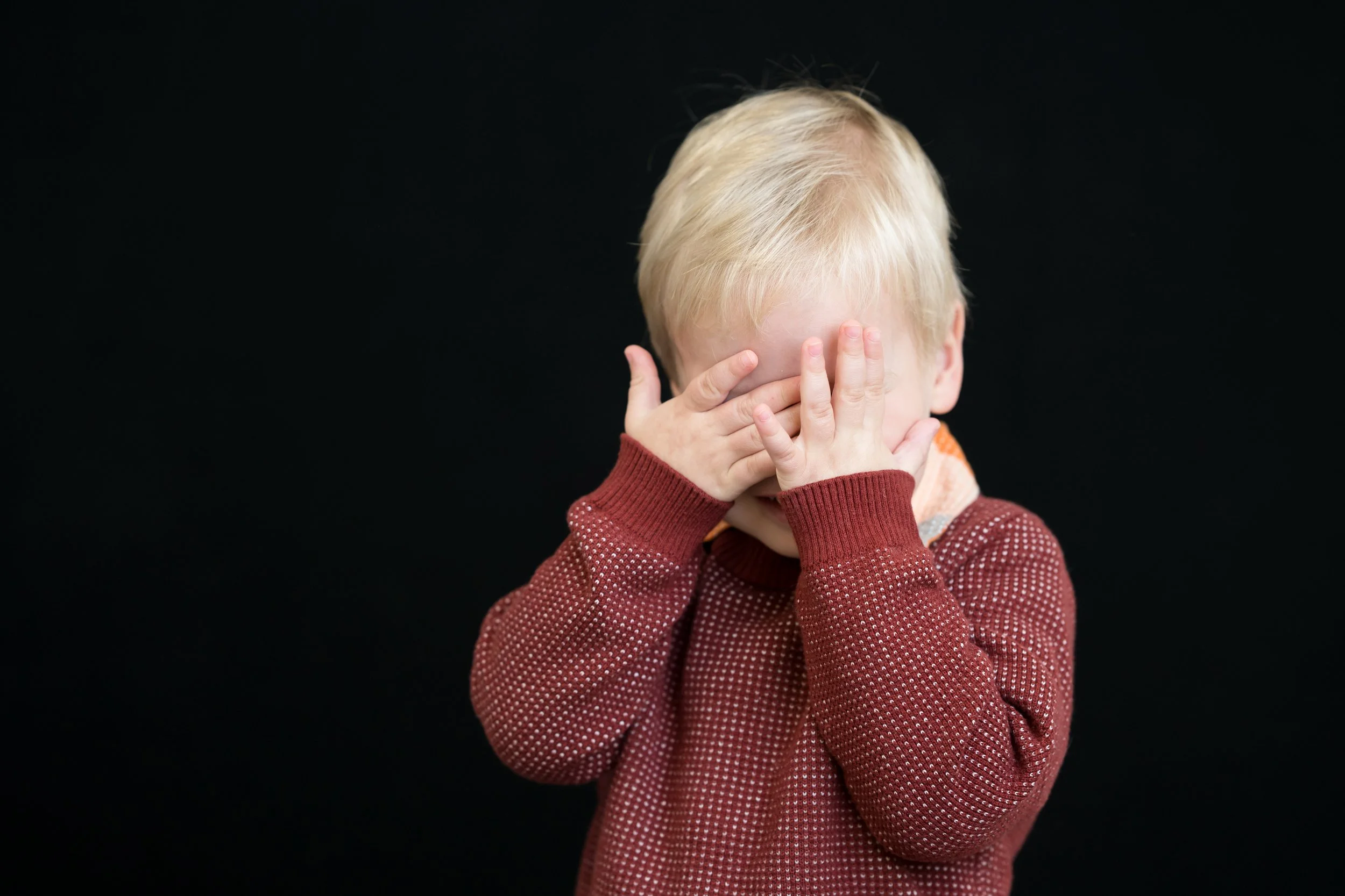 A young child with blond hair covering their face with both hands, wearing a textured maroon sweater, against a black background.