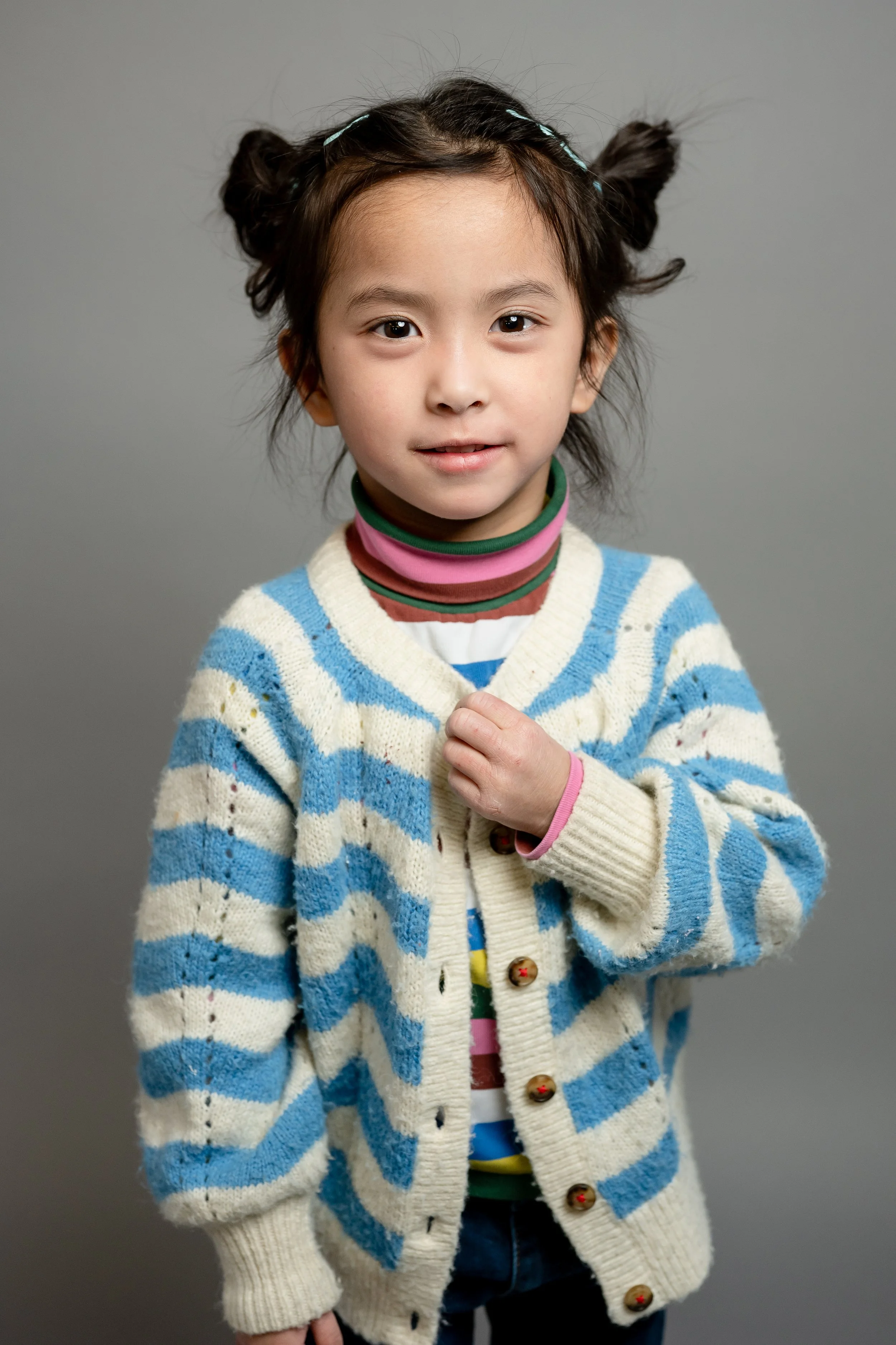 A young girl with dark hair styled in two buns, wearing a cream and blue striped sweater, layered shirts with colorful stripes, and a pink and green turtleneck, standing against a gray background.