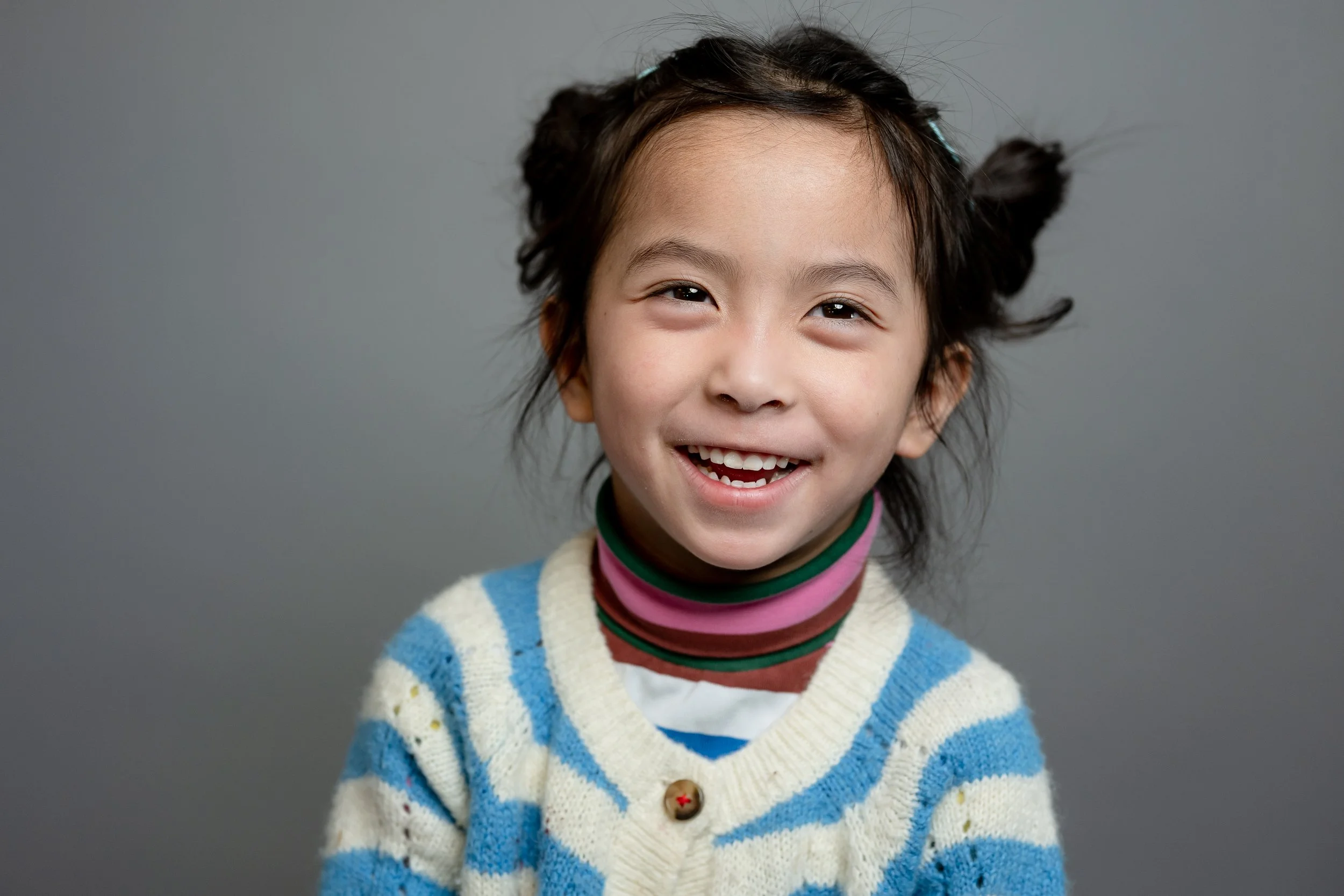 A young girl with dark hair styled in two messy buns, smiling and showing her teeth, wearing a striped cream and blue sweater and a multicolored turtleneck, against a plain gray background.