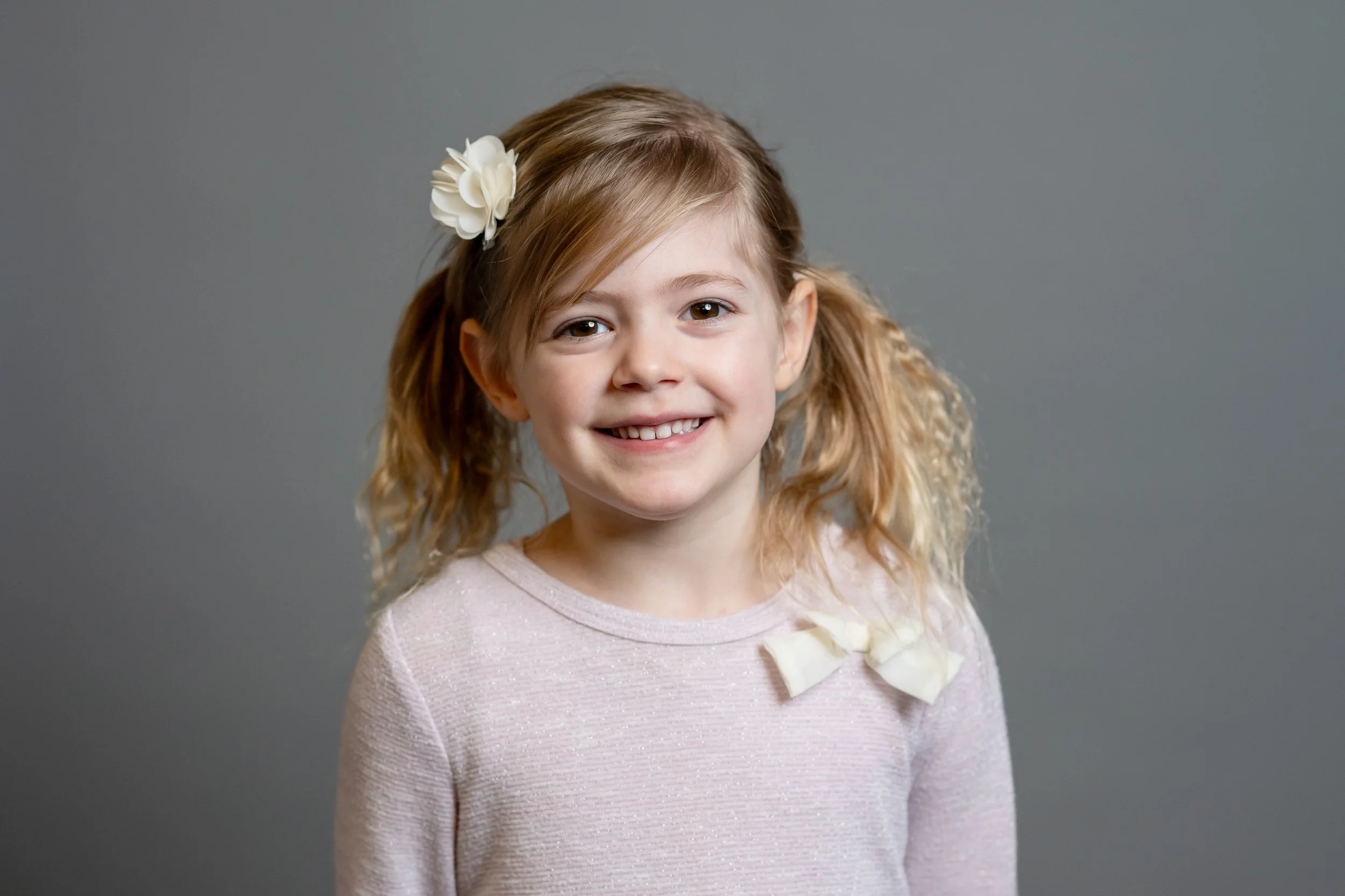 A smiling young girl with light brown hair tied in pigtails, wearing a light pink sweater with a bow on the collar and a white flower hair clip, standing against a gray background.