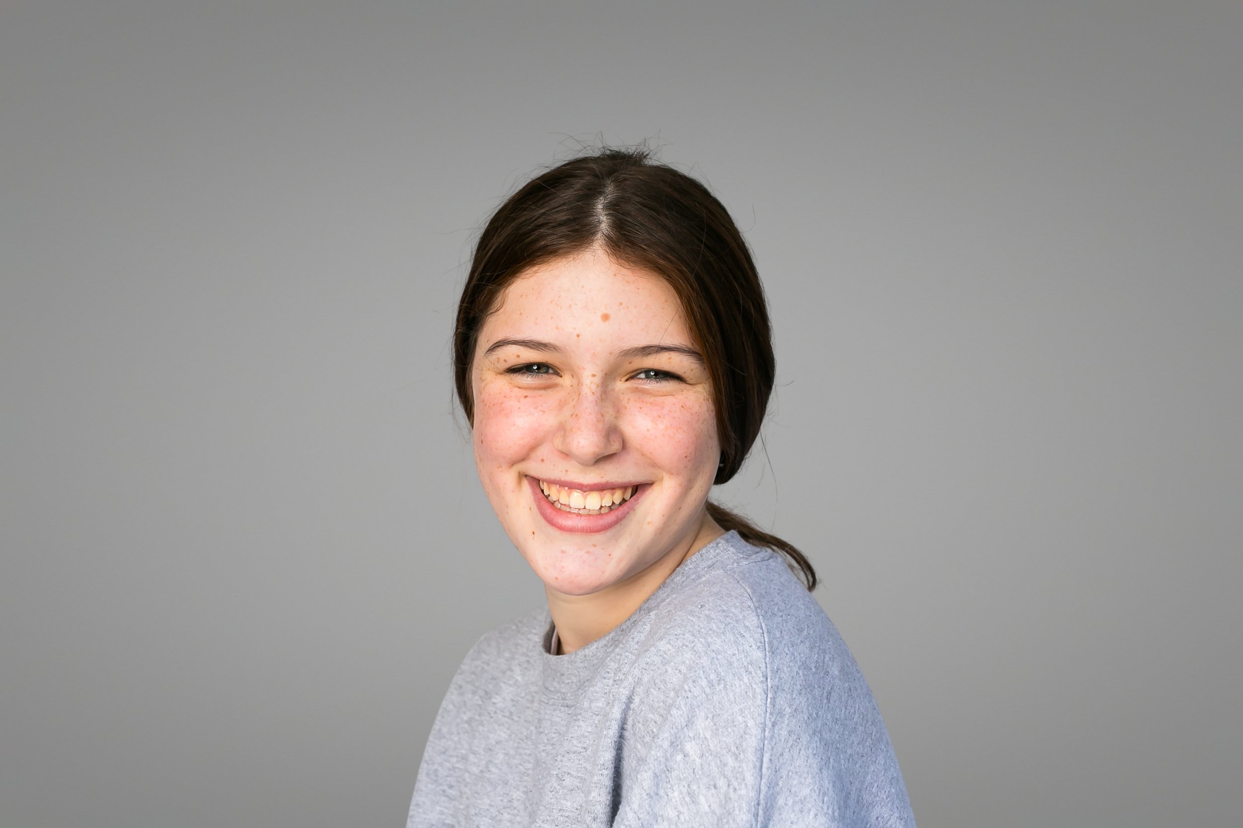 Smiling young woman with dark hair and freckles wearing a light gray shirt against a plain gray background.