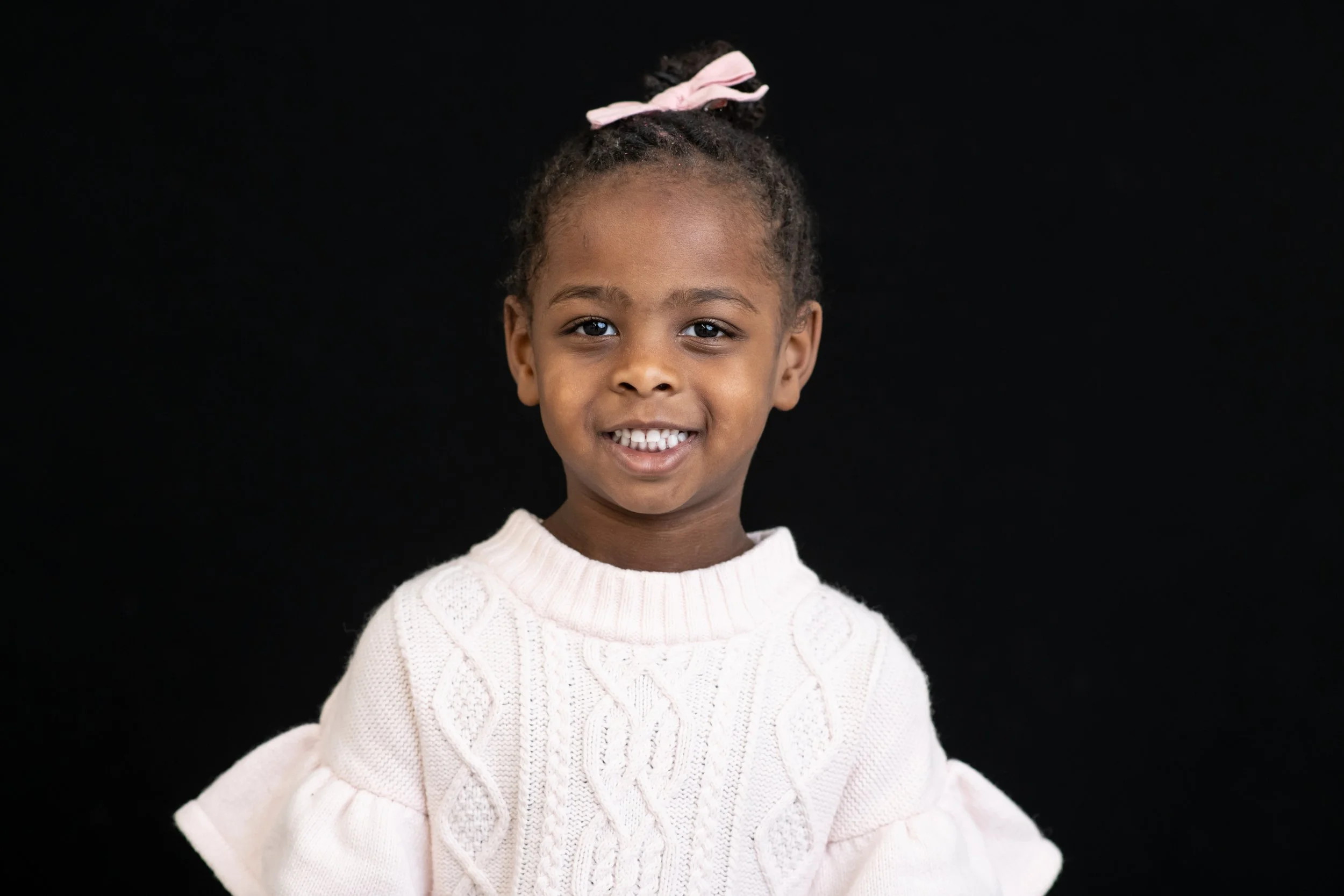Close-up portrait of a young girl with braided hair and a pink bow, smiling, wearing a white knit sweater, against a black background.