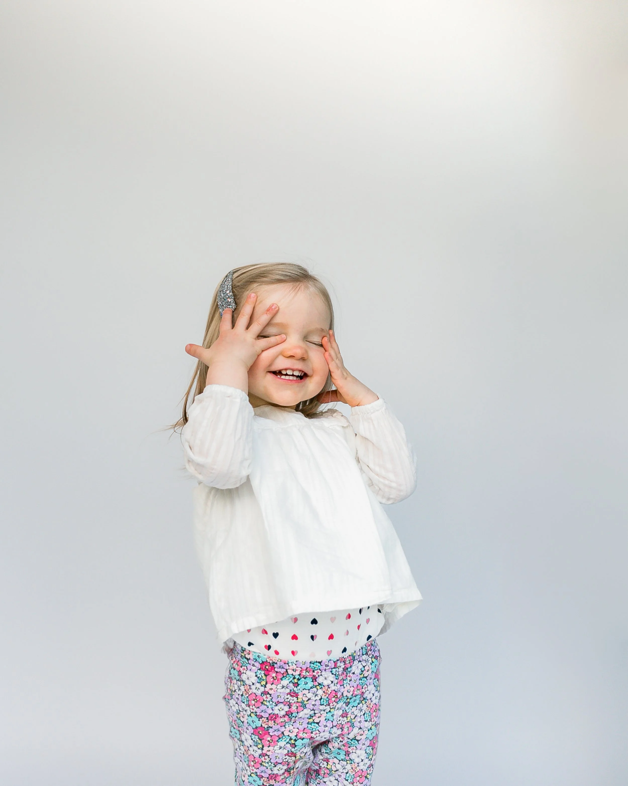 A young girl with a big smile covering her eyes and face with her hands, wearing a white long-sleeve shirt, floral pants, and a glittery hair clip, standing against a plain white background.