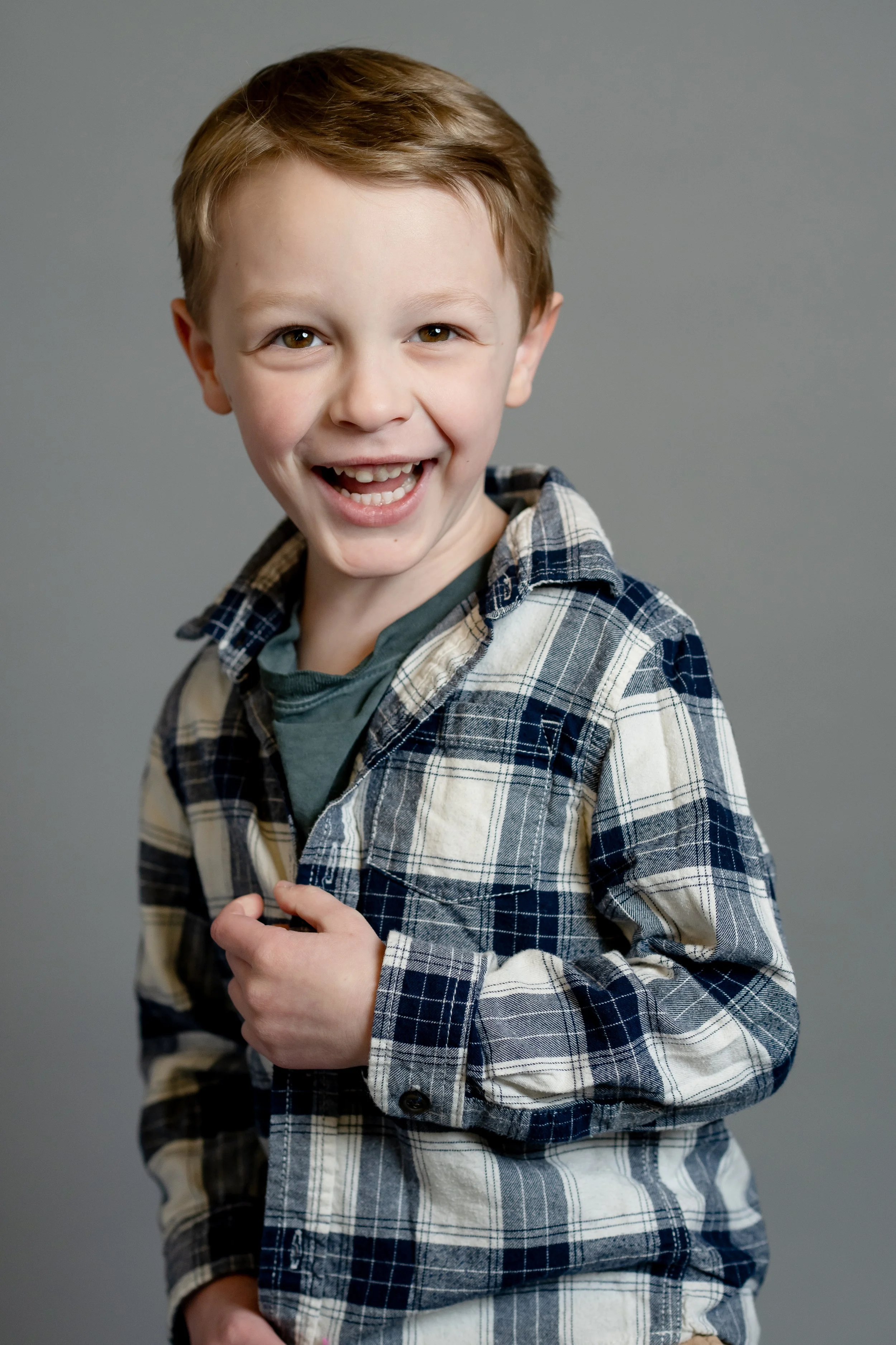 A young boy with brown hair smiling and showing his teeth, wearing a blue and white plaid shirt over a gray T-shirt, against a plain gray background.