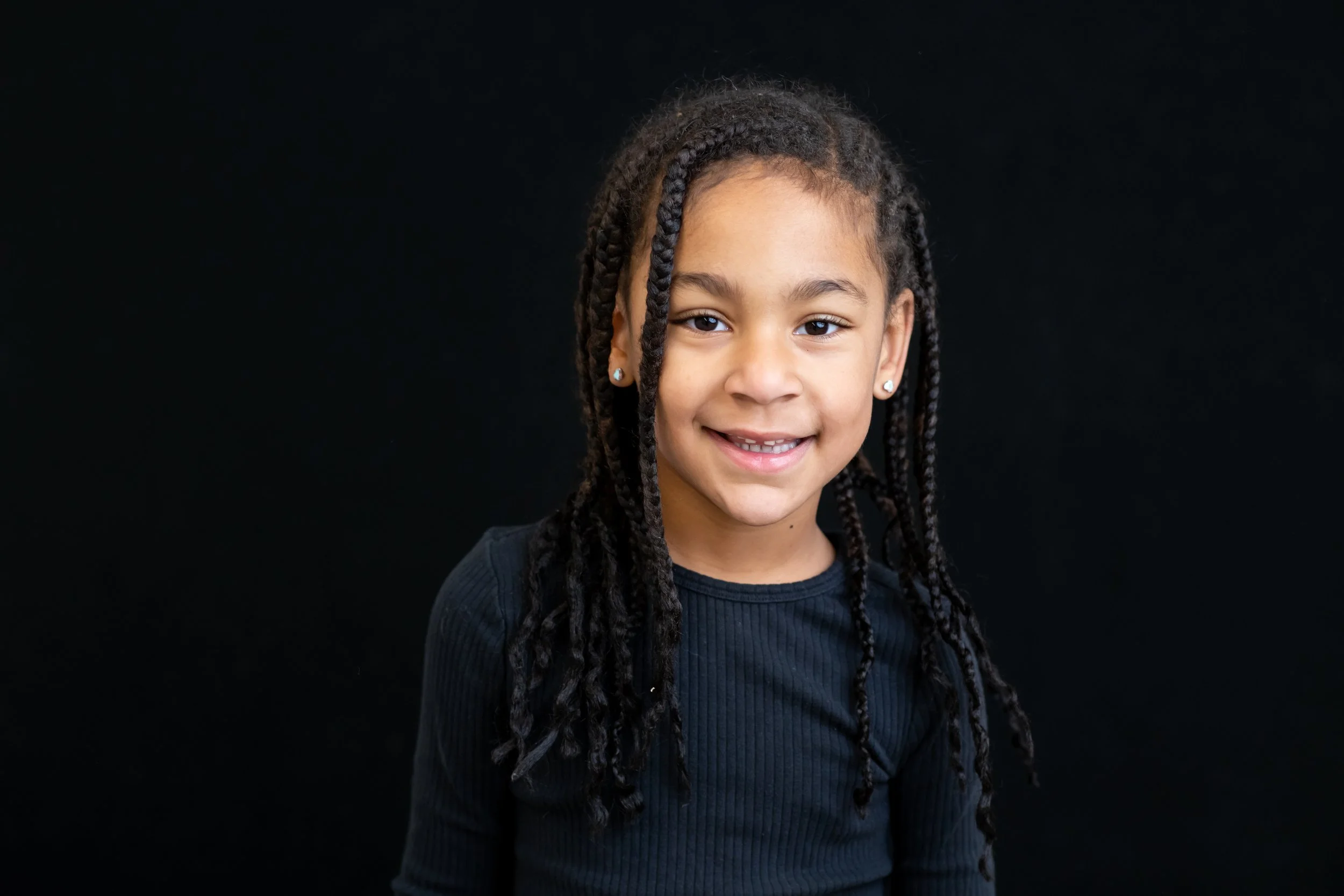 Portrait of a young girl with braids, smiling, wearing a black top, against a black background.