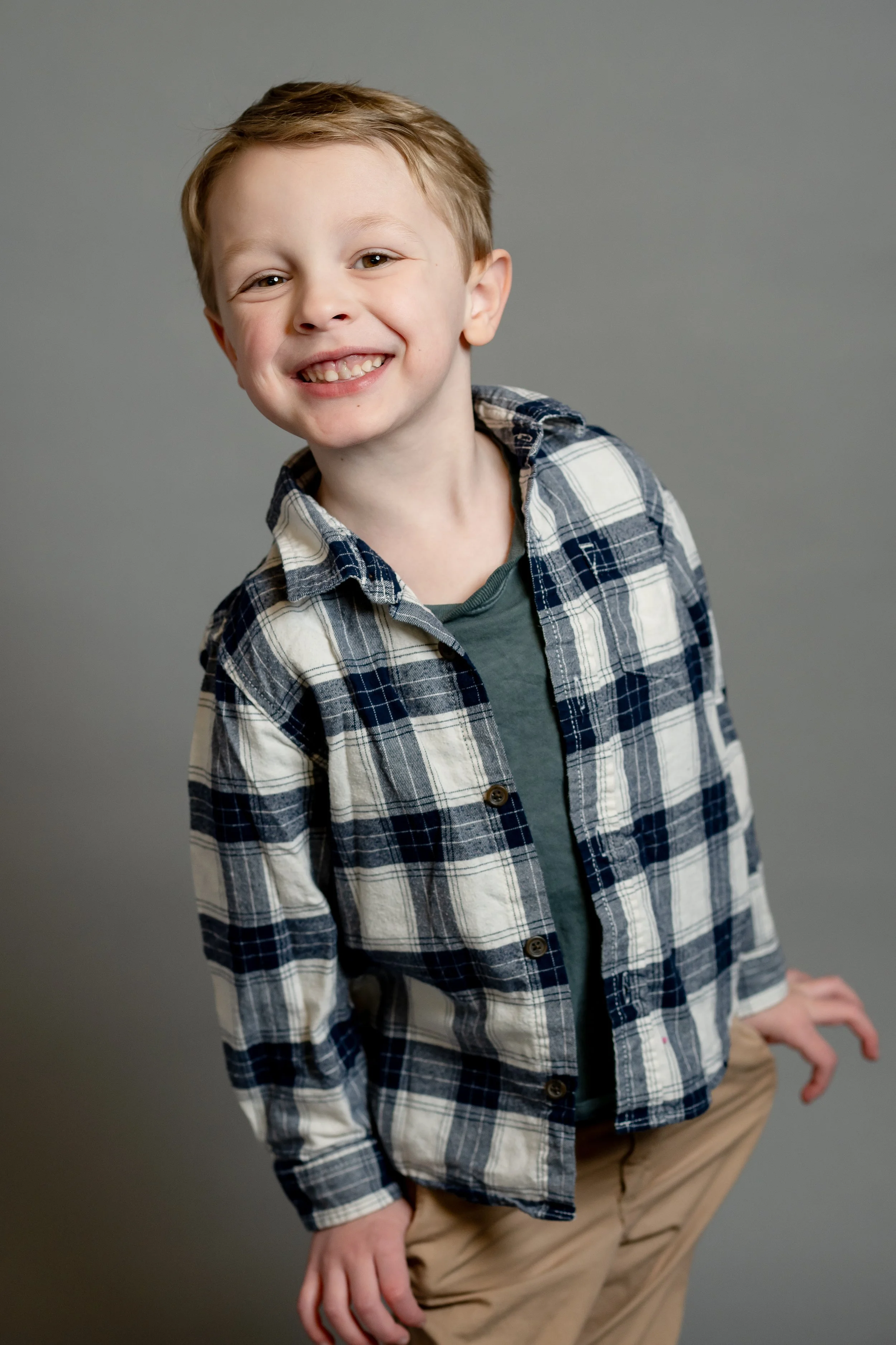 Smiling young boy with light brown hair, wearing a blue and white plaid shirt over a green T-shirt, standing against a gray background.