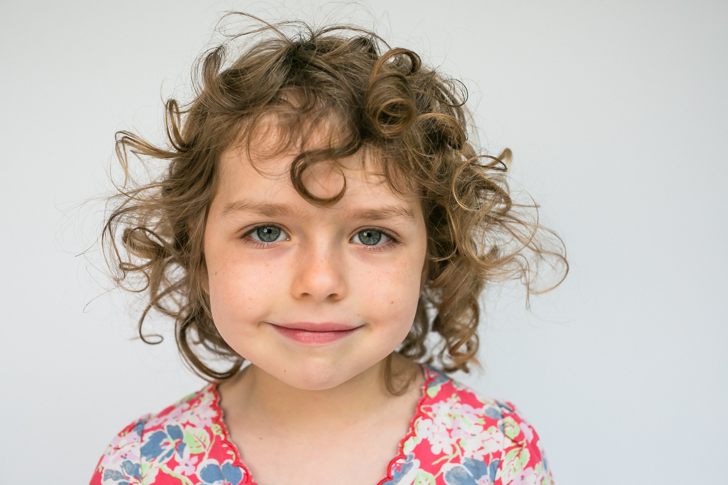 Close-up of a young girl with curly brown hair and blue eyes, wearing a colorful floral top, smiling slightly against a plain white background.