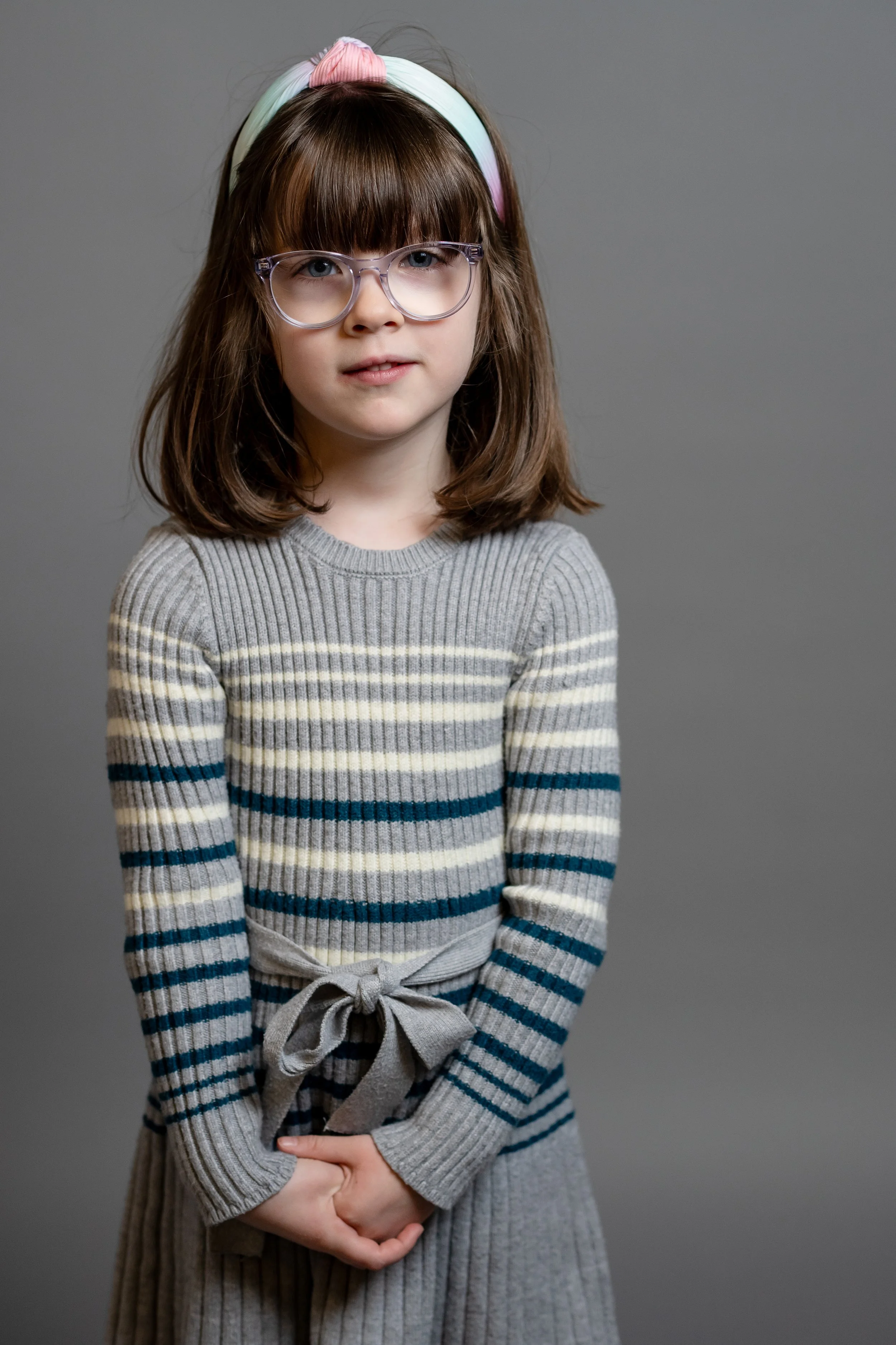 A young girl with brown hair, glasses, and a pastel headband stands against a plain gray background, wearing a gray striped sweater and skirt.