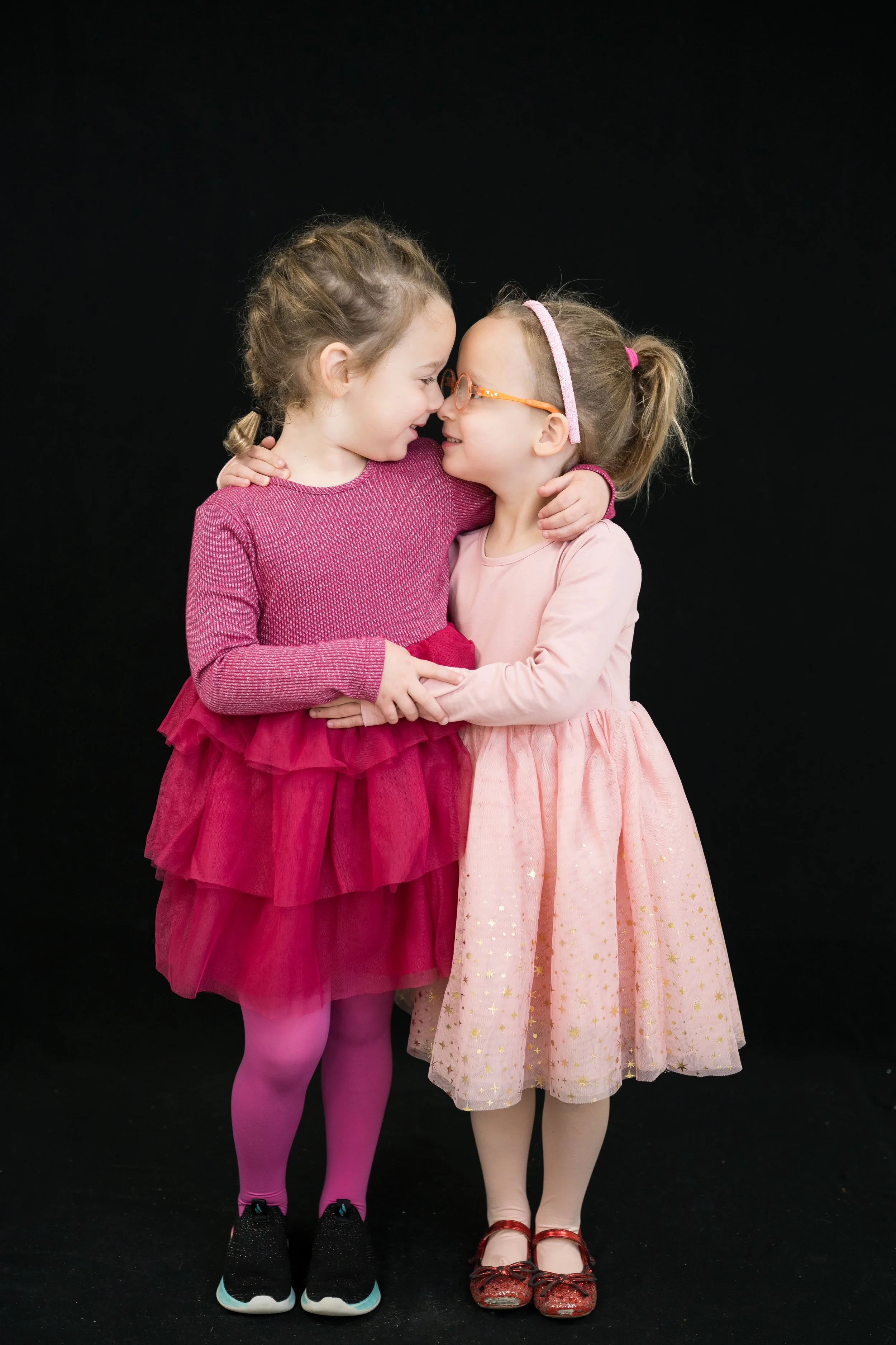 Two young girls in pink dresses hugging and touching noses, smiling against a black background.