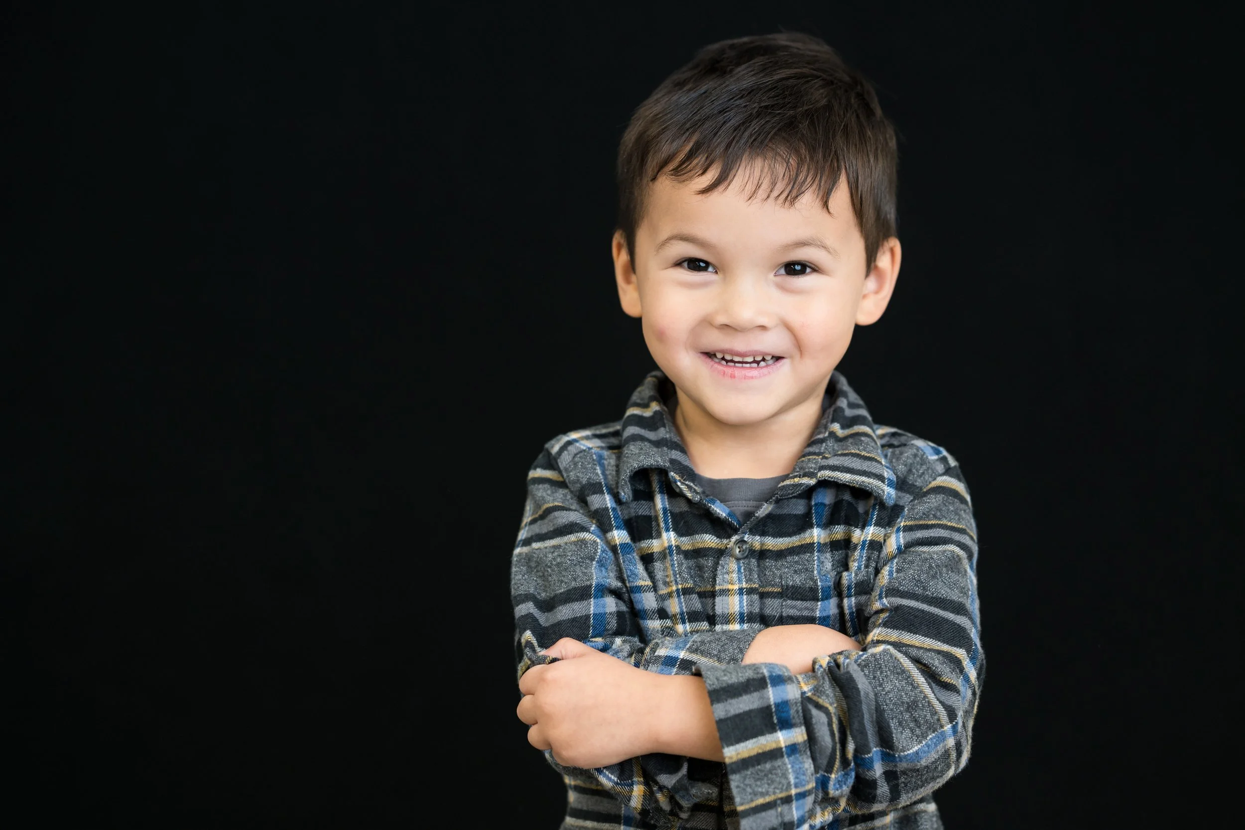 A young boy with dark hair smiling and crossing his arms, wearing a plaid shirt, against a black background.