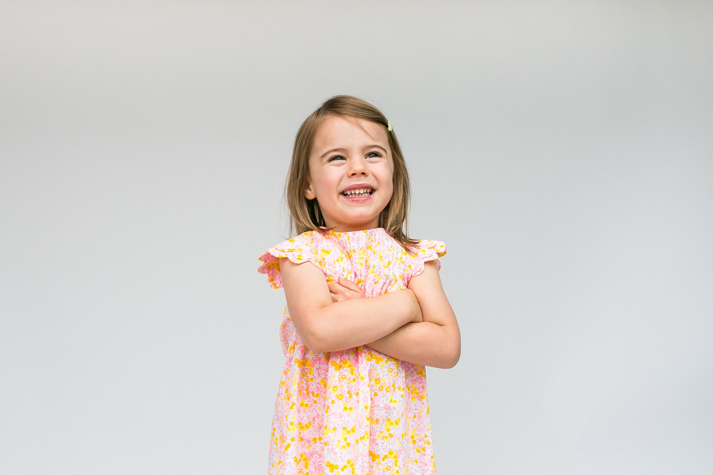 A young girl with brown hair, smiling and crossing her arms, wearing a pink floral dress, standing against a plain light gray background.