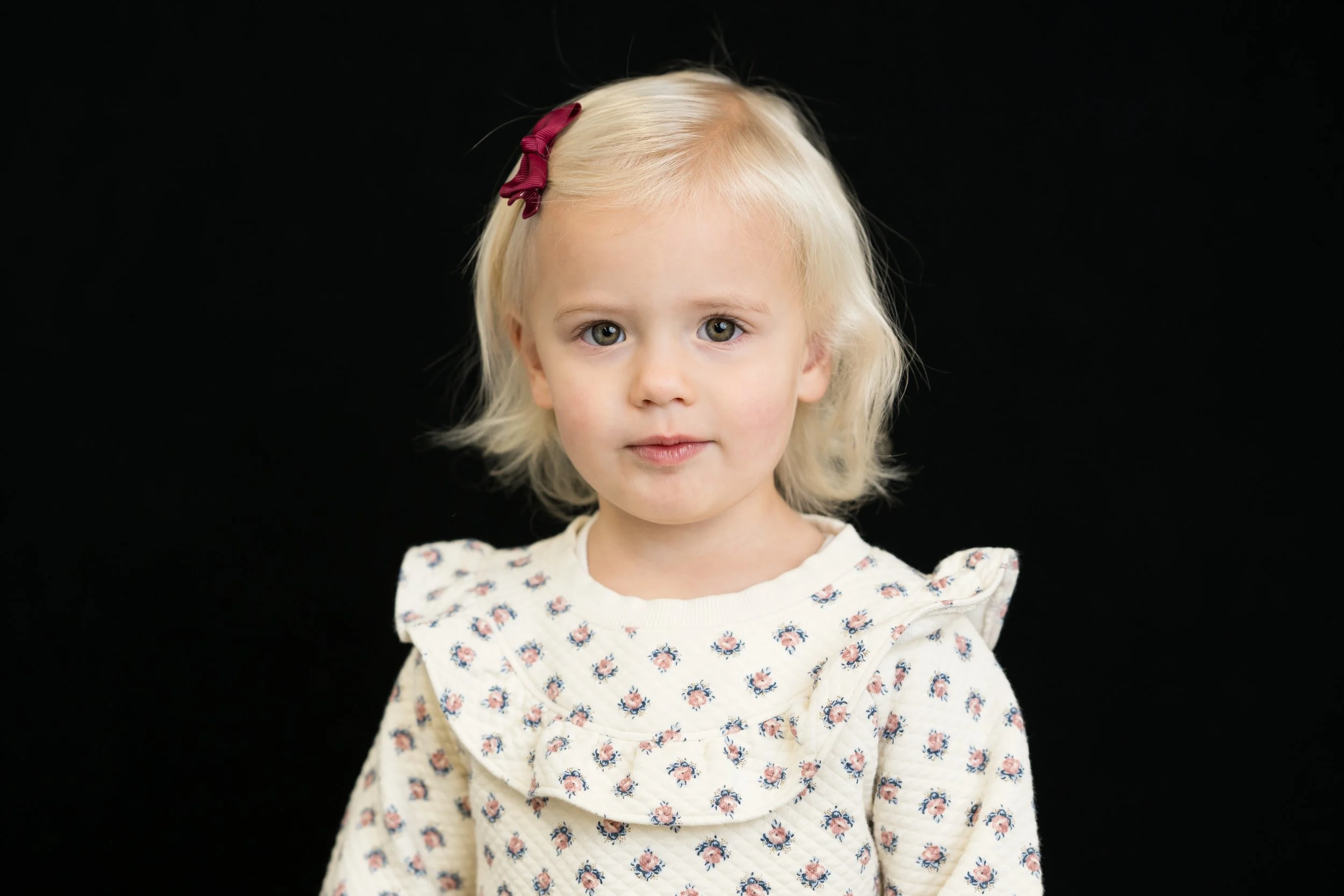 A young girl with blonde hair, wearing a cream-colored sweater with a floral pattern and a dark red hair clip, standing against a black background.