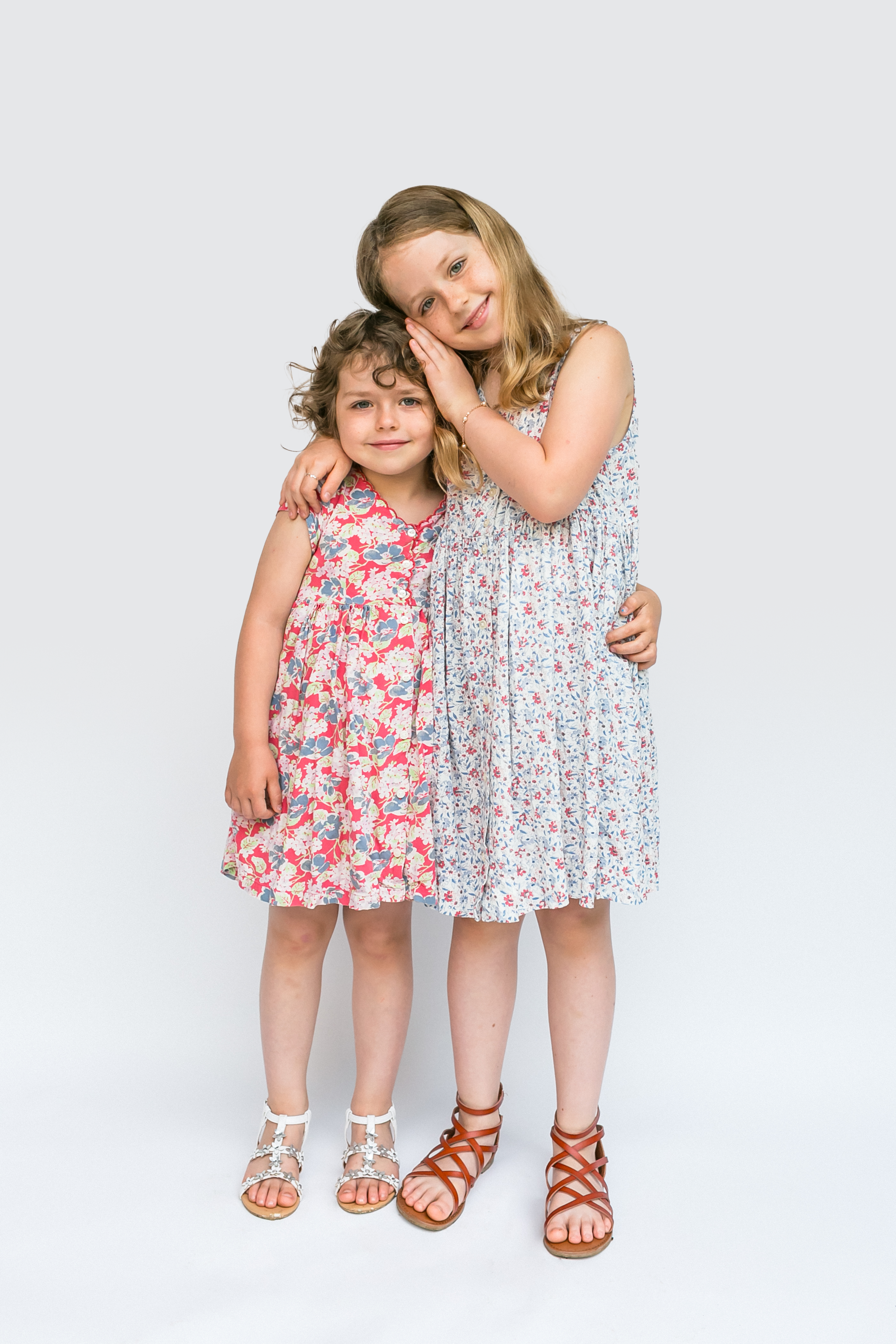 Two young girls standing close together, one with her head leaning on the other's cheek, both wearing floral dresses, smiling against a plain white background.