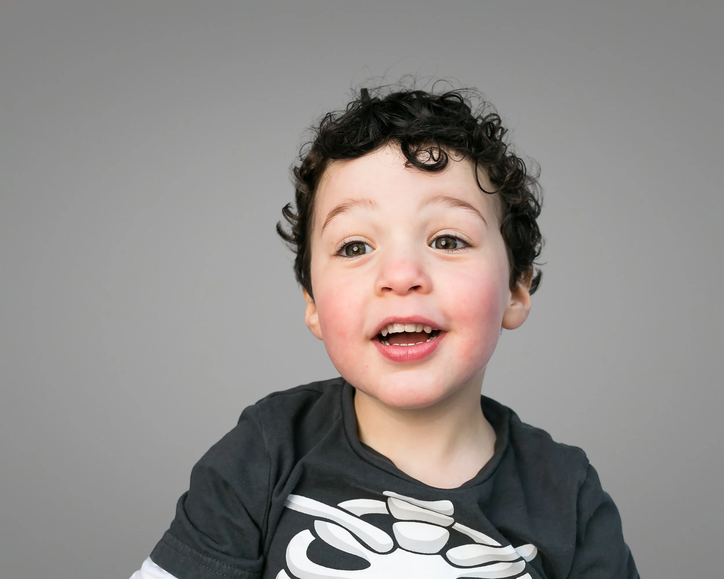 Close-up of a young boy with curly hair, smiling, wearing a black t-shirt with a white skeletal design, against a plain gray background.