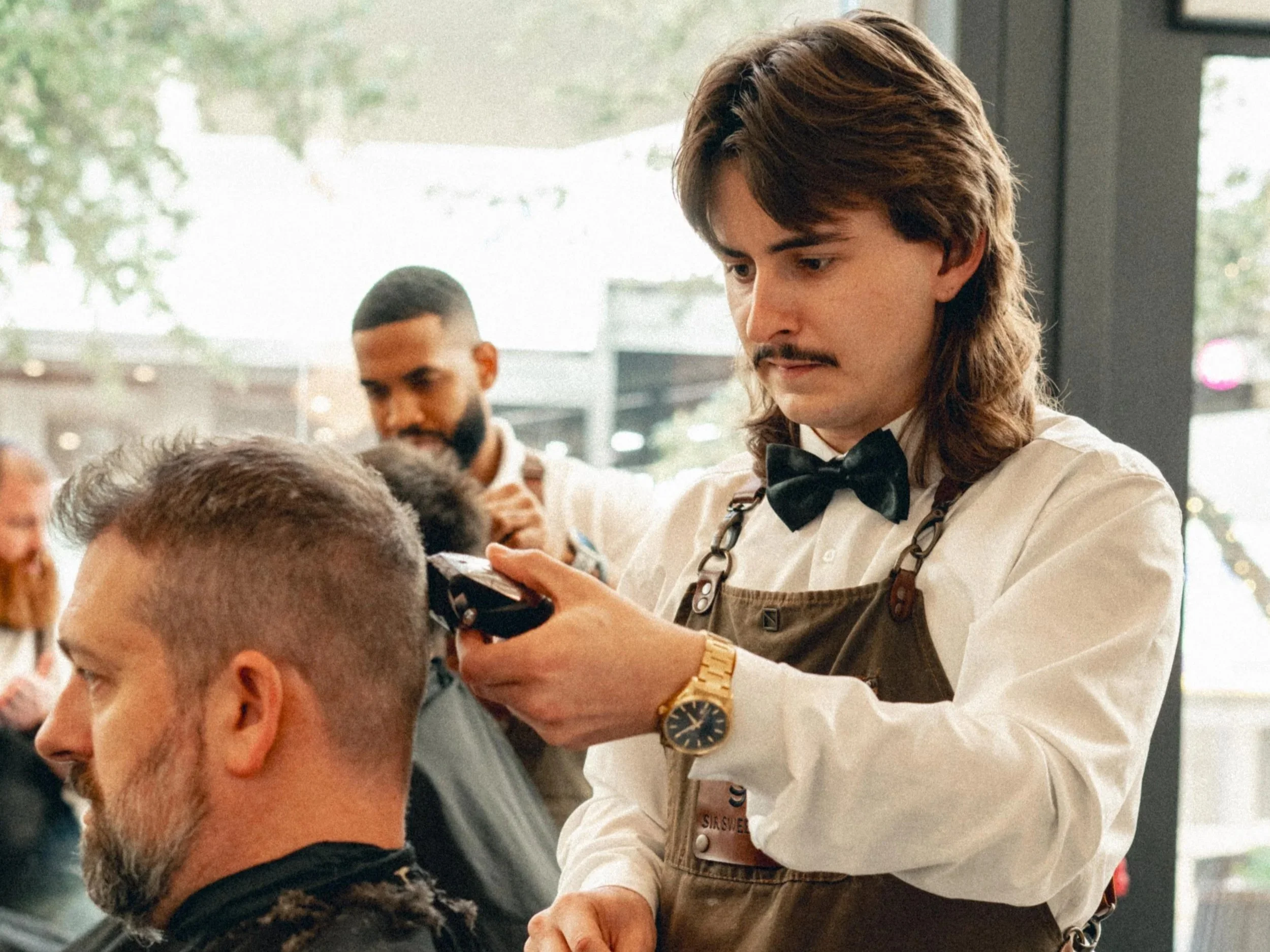 Barber in apron giving haircut to a man with glasses and beard while another barber cuts hair in the background inside a barbershop.