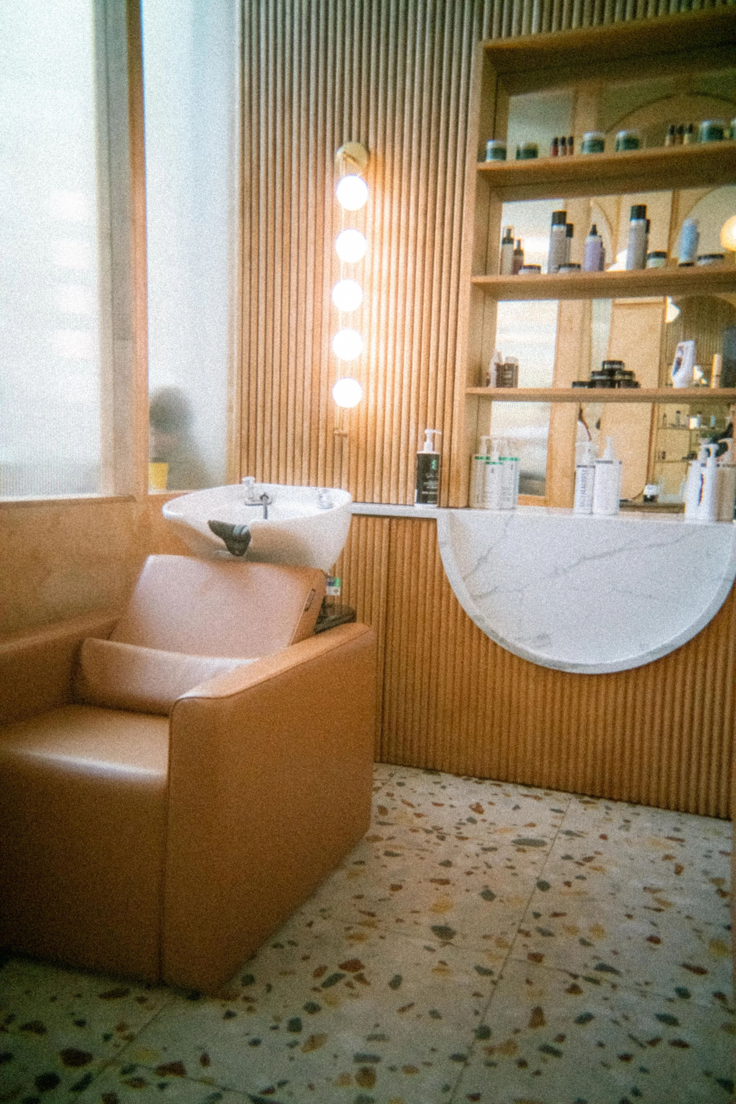 A hair salon or spa station with a tan hair washing chair, a sink, and wooden shelves with hair products. The wall is lined with vertical wooden slats, and there are four round lights hanging on the wall.