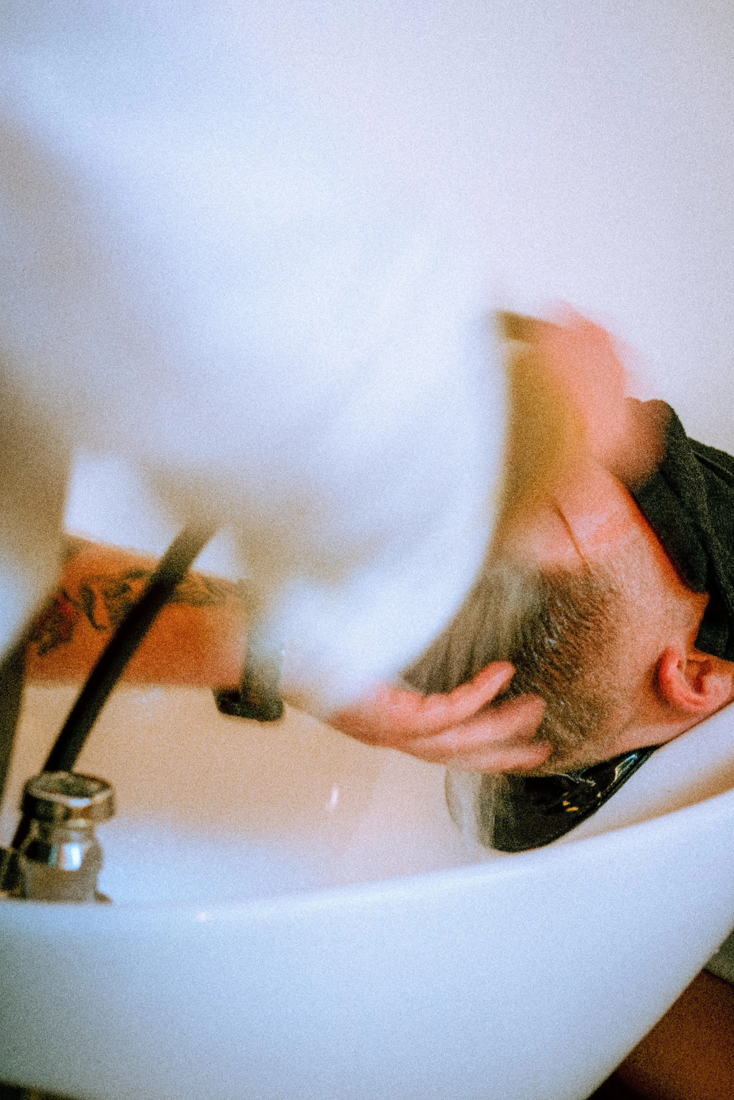 A man receiving a haircut at a salon, leaning over a white sink with a black hose attached, while the barber cuts his hair.