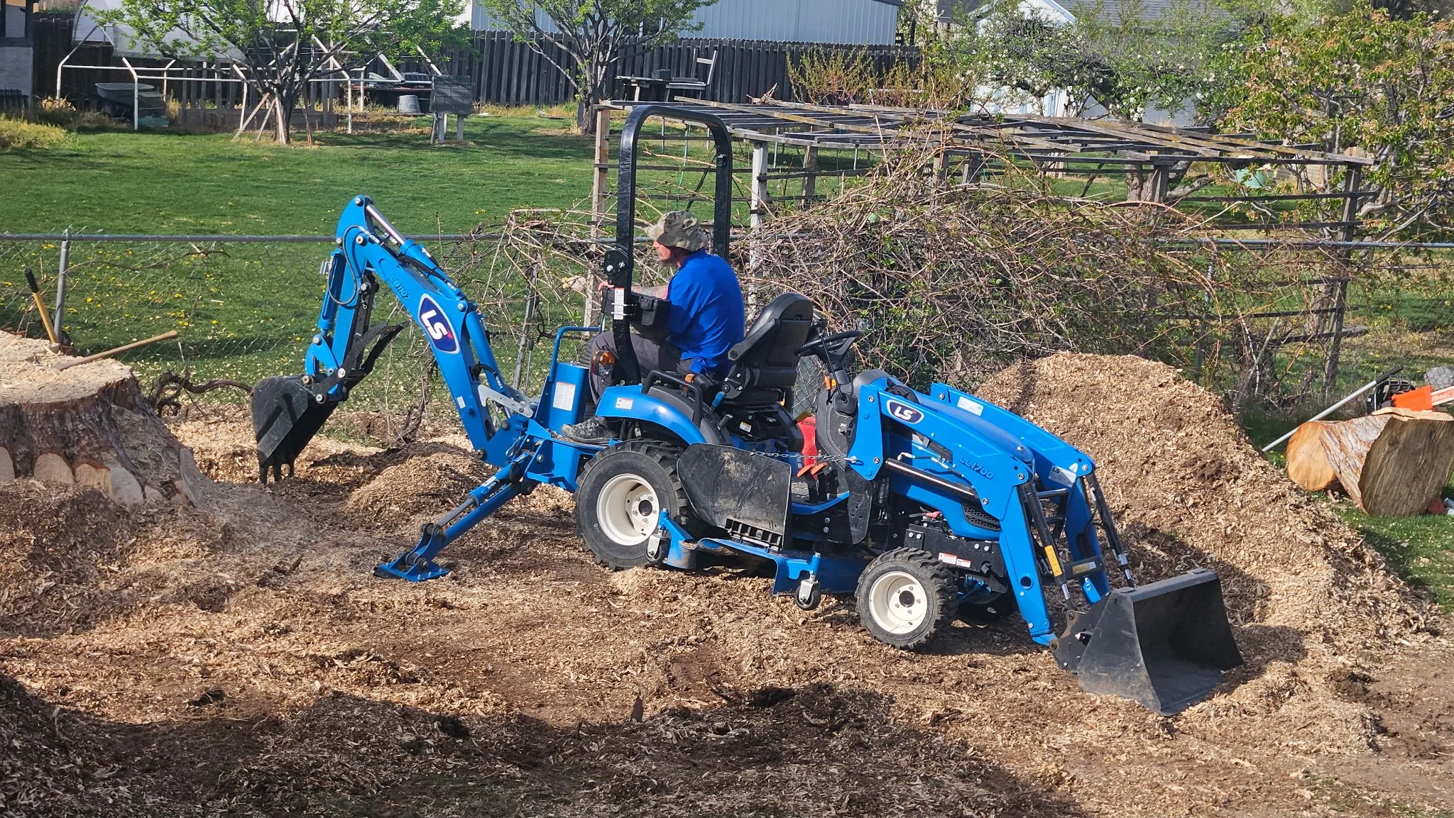 A person operating a blue compact excavator on a dirt surface, with fallen tree branches and logs, in a backyard with green grass, trees, and a fenced area.