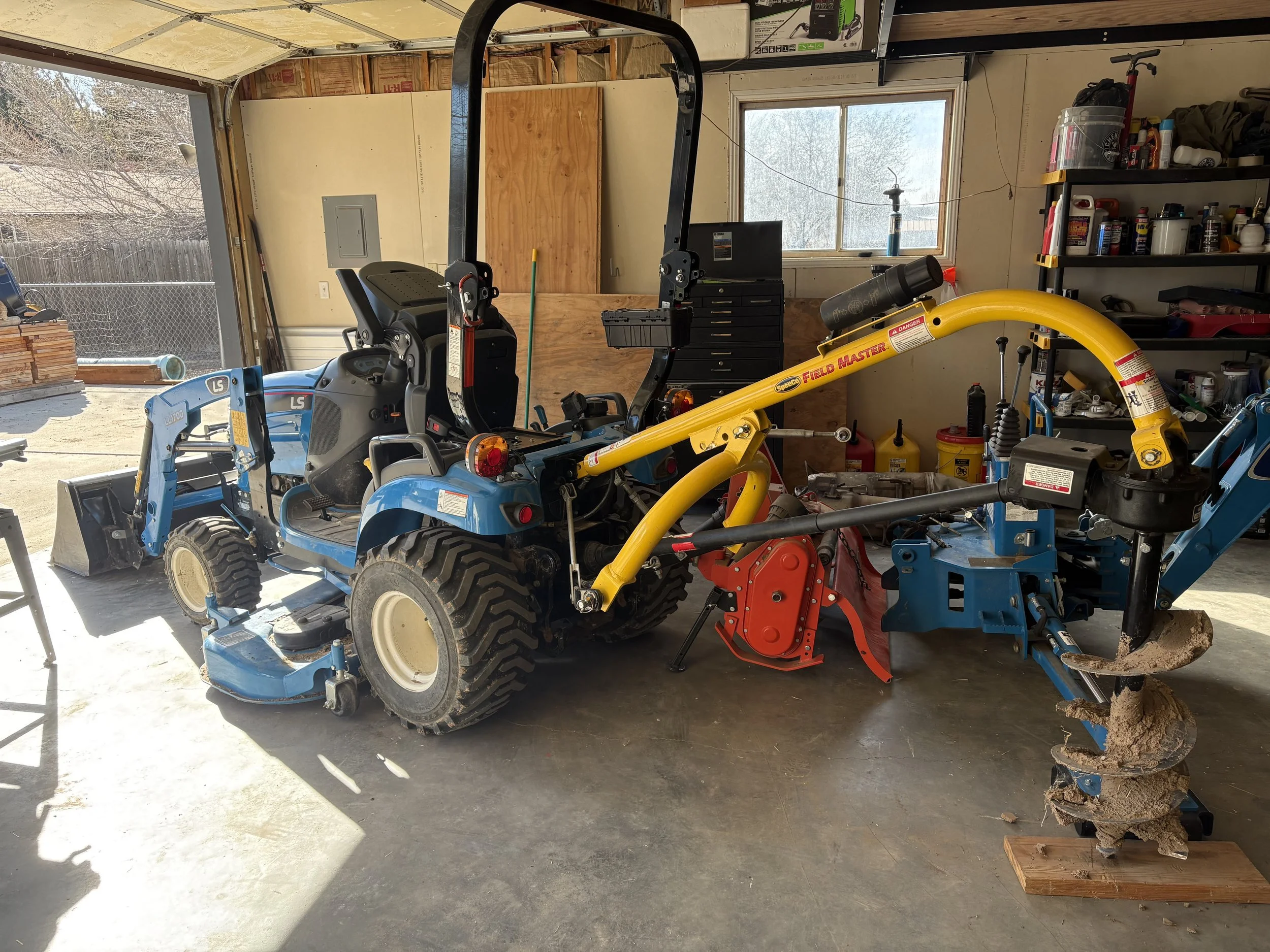A garage with a blue tractor, yellow equipment, and shelves with bottles and tools.