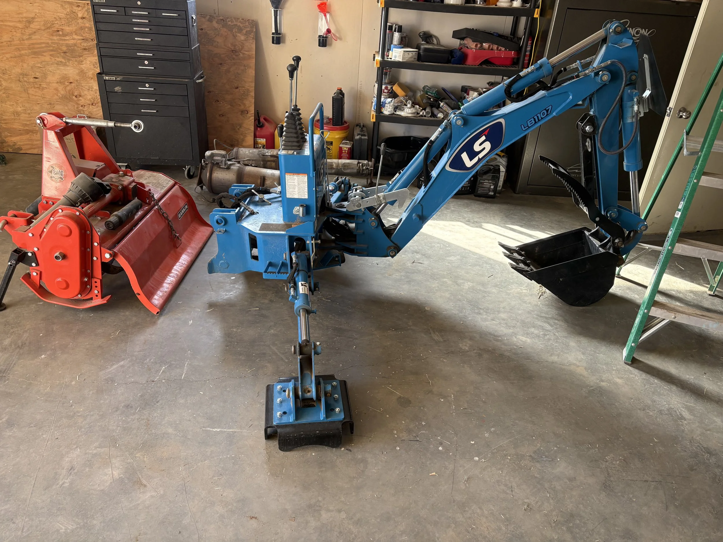 A garage workshop with a blue mini excavator and a red attachment, black tool chest, shelving with various tools and supplies, and a ladder. The floor is concrete with some marks and debris.