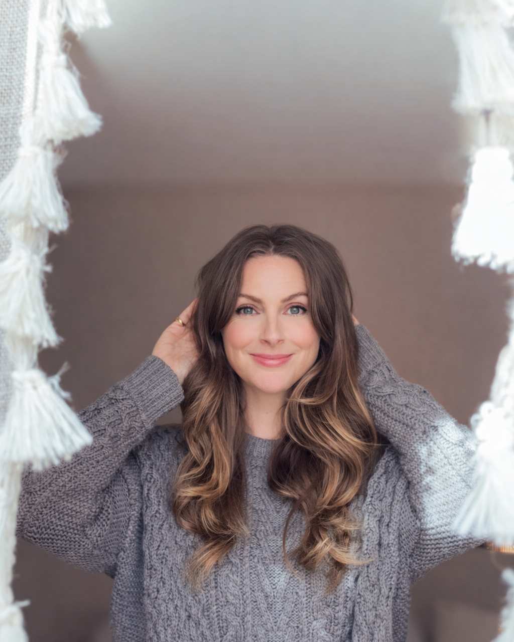 A woman with long, wavy brown hair and blue eyes smiling while looking into the camera, framing her face with her hands behind her head, wearing a gray knit sweater.