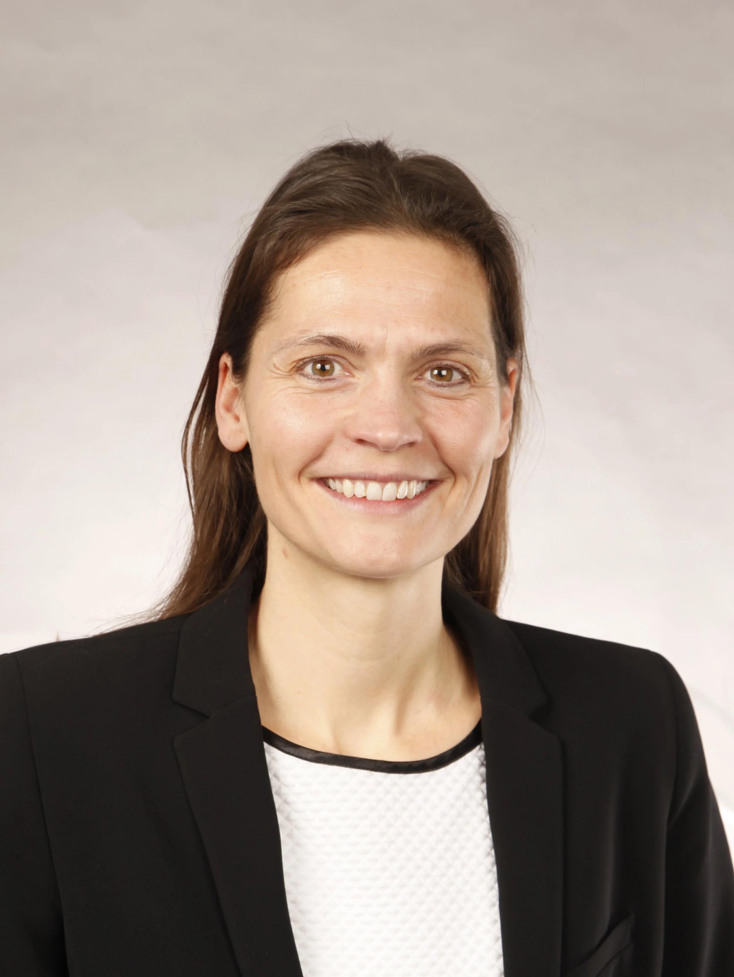 Porträt einer lächelnden Frau mit braunen Haaren, im schwarzen Blazer und einer weißen Bluse, vor hellem neutralem Hintergrund.