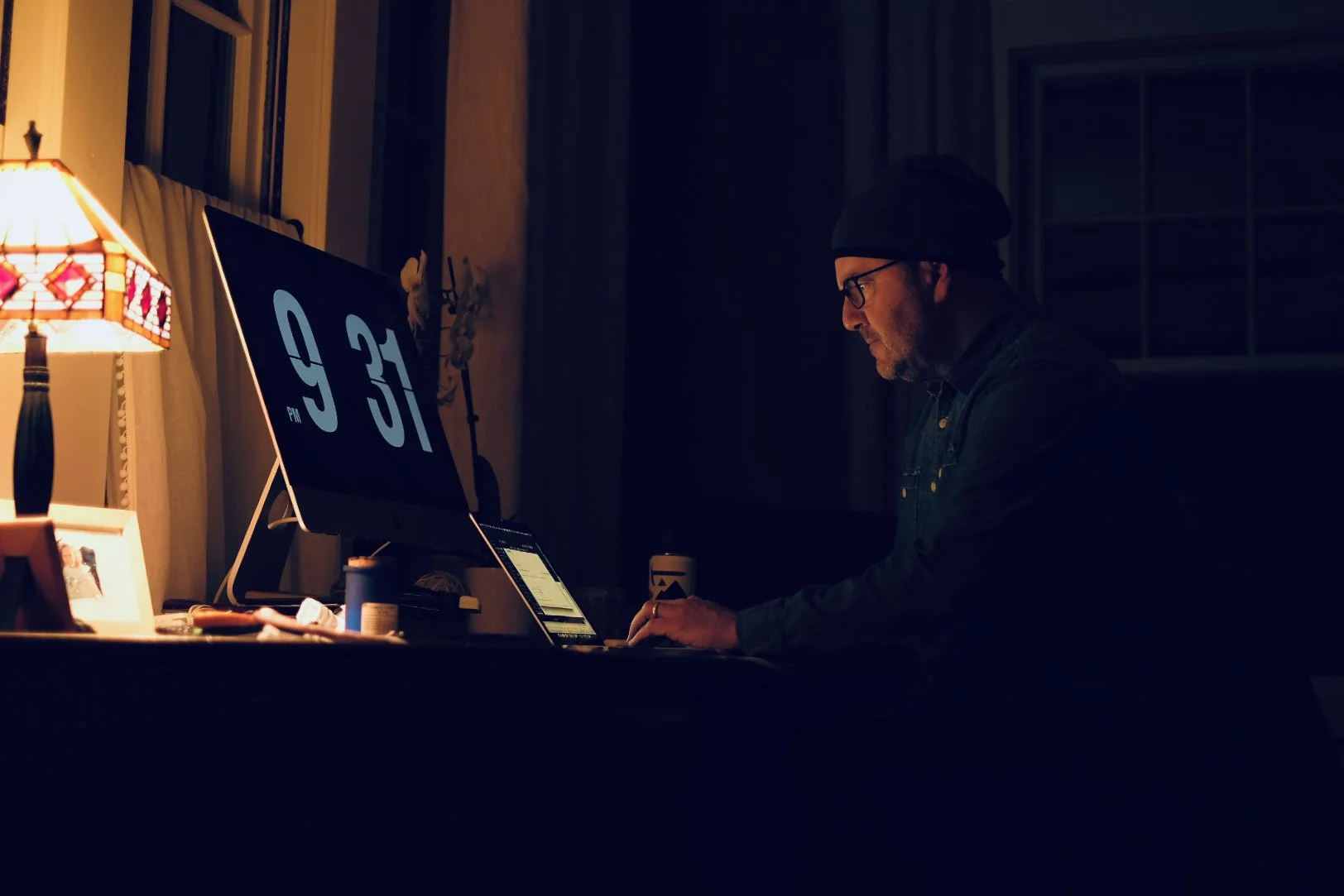 A man wearing glasses, a beanie, and a dark jacket working at a desk in a dimly lit room with a computer monitor displaying the time 9:31.