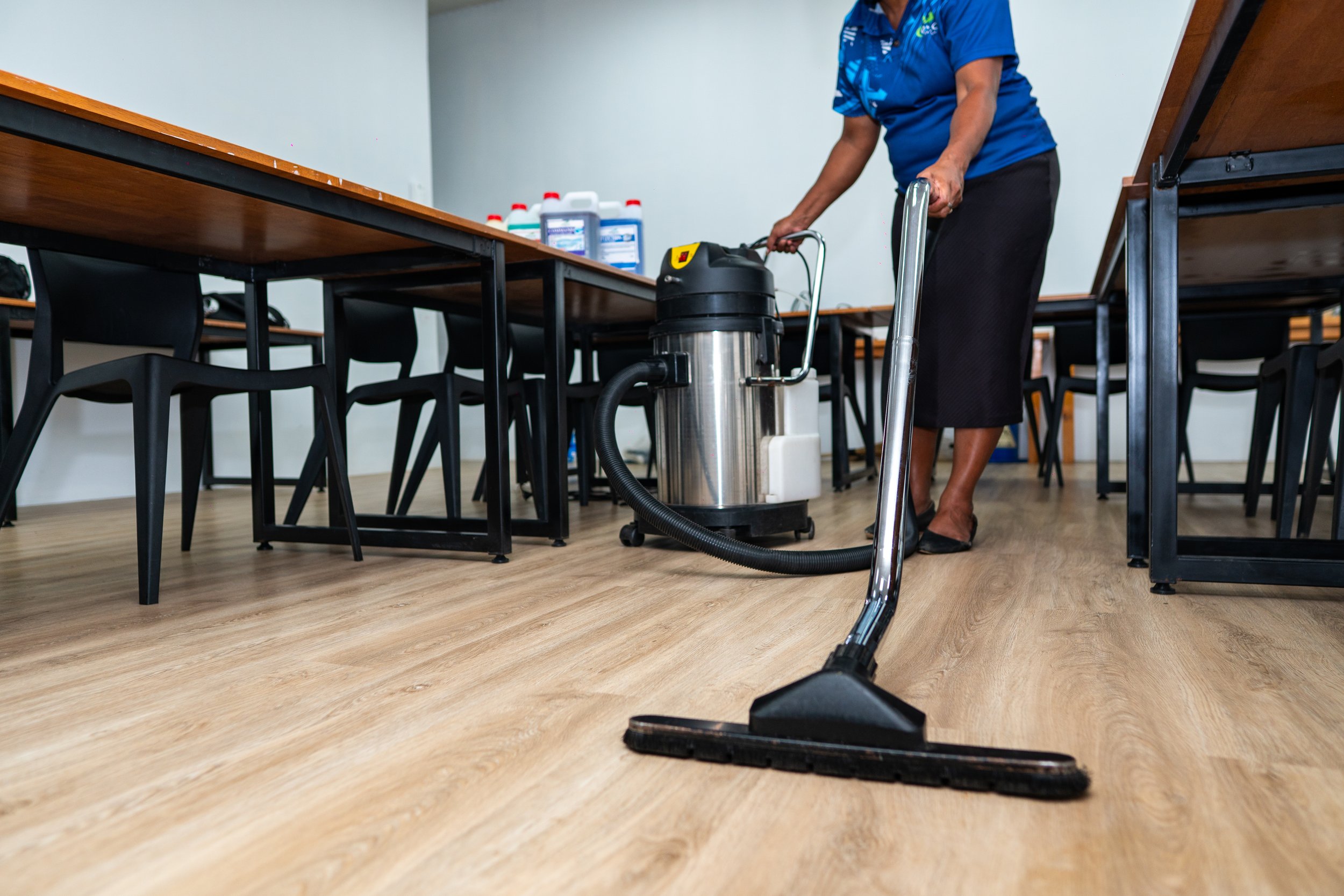 A person in protective gear, including a full-body suit, gloves, mask, and goggles, is using a fogging machine to disinfect a carpeted floor indoors.