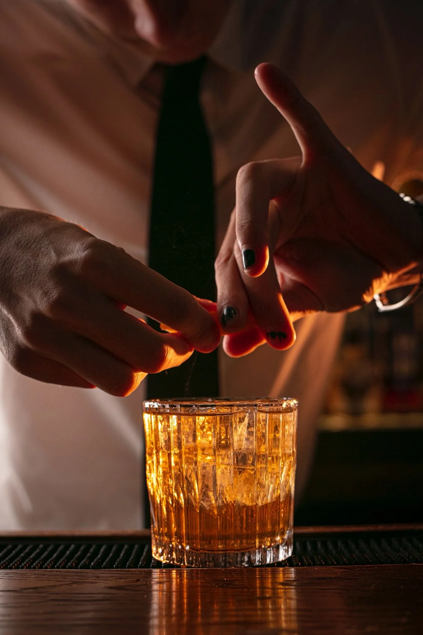 A bartender prepares an amber-colored cocktail in a textured glass, using a script or garnish held with both hands. The setting is warm and dimly lit.
