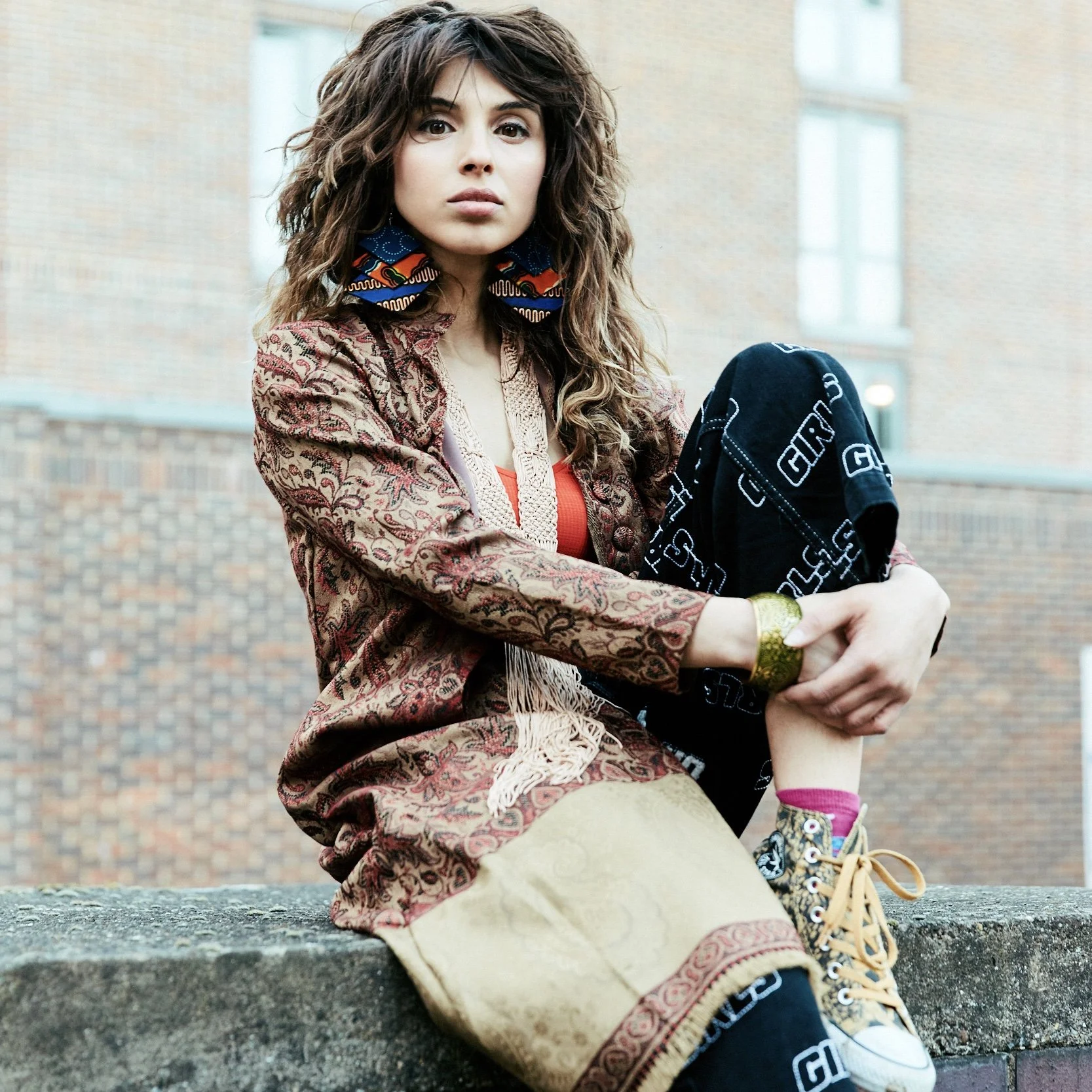 A young woman with long, curly brown hair sitting on a concrete ledge outdoors, wearing colorful and eclectic clothing including patterned sneakers, a patterned jacket, and large earrings, with a brick building in the background.