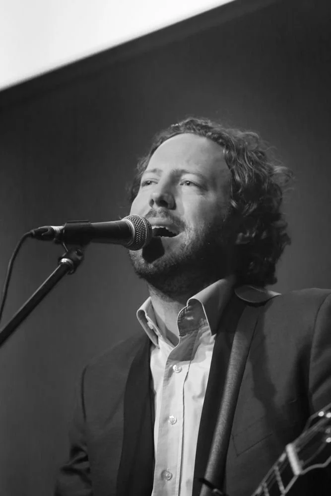 A man with curly hair singing into a microphone while playing a guitar, dressed in a suit and shirt.
