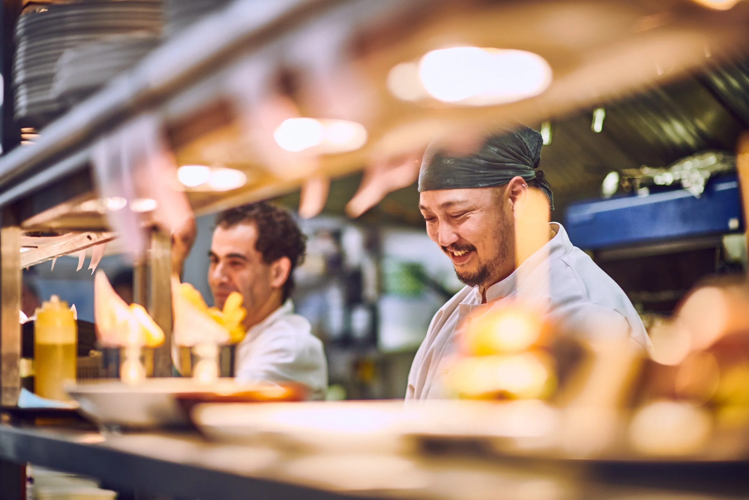 Two chefs working in a restaurant kitchen, smiling as they prepare food behind a counter with condiments and cooking supplies.