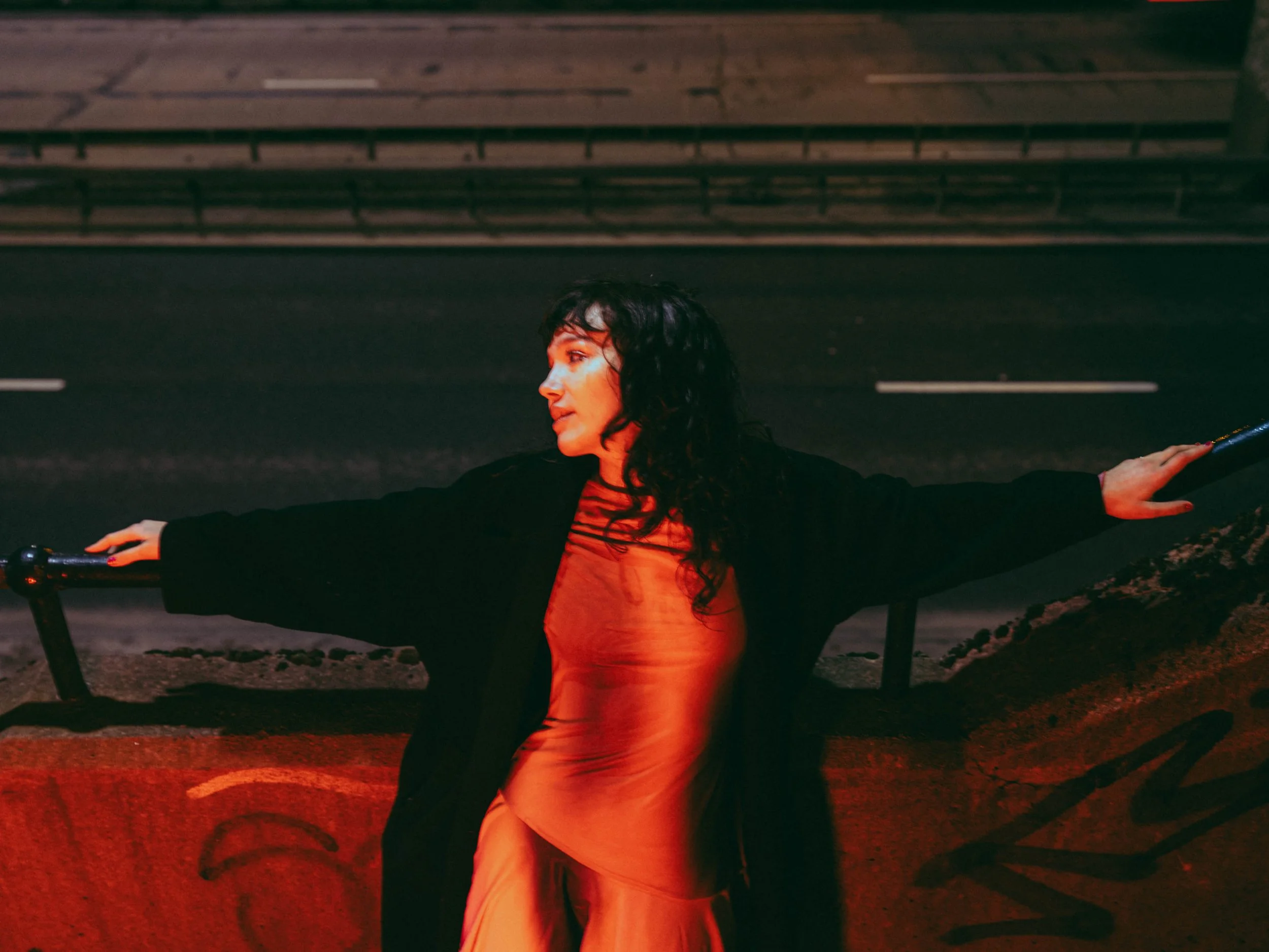 A woman with dark, curly hair standing with arms outstretched, leaning against a black railing at night, with a dark background and red lighting illuminating her face and dress.
