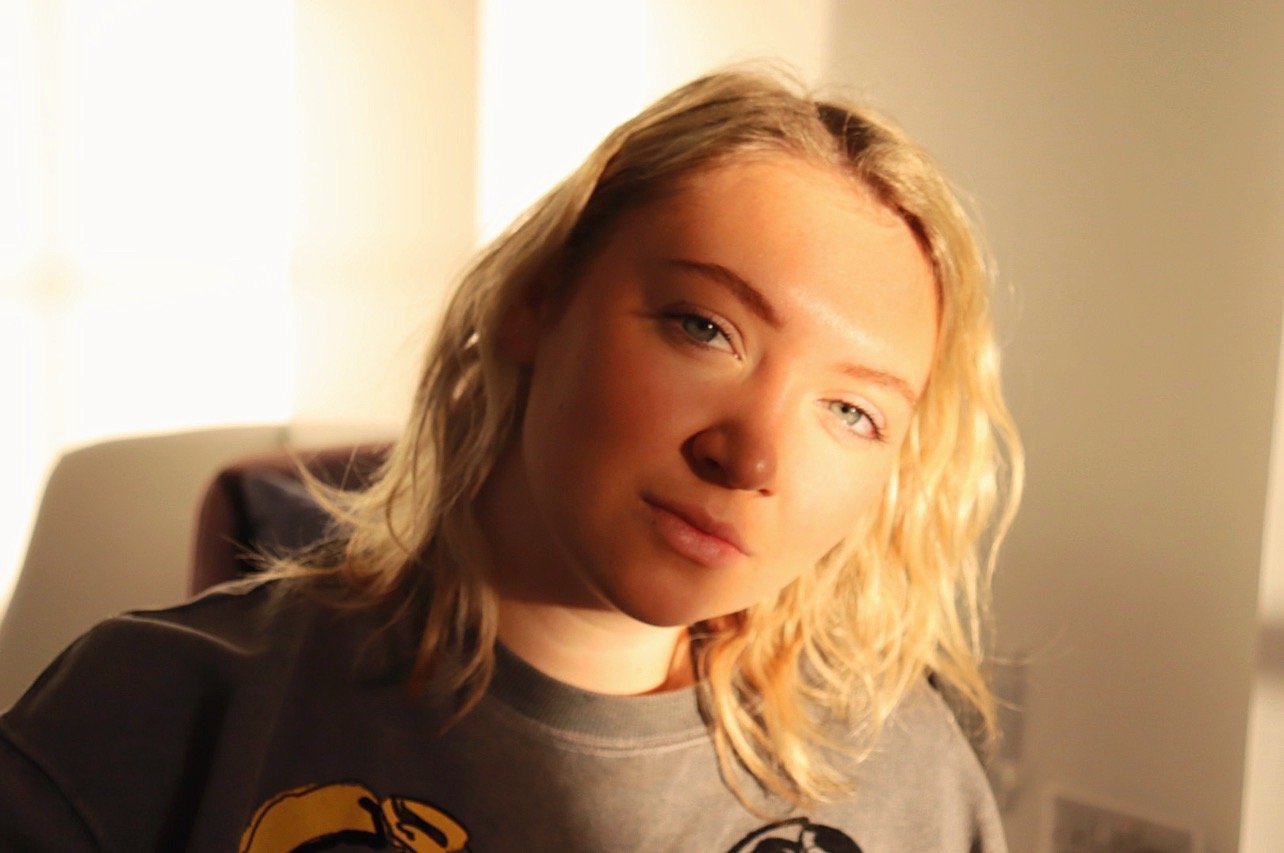 A young woman with blonde wavy hair and blue eyes looking at the camera in a warmly lit room.