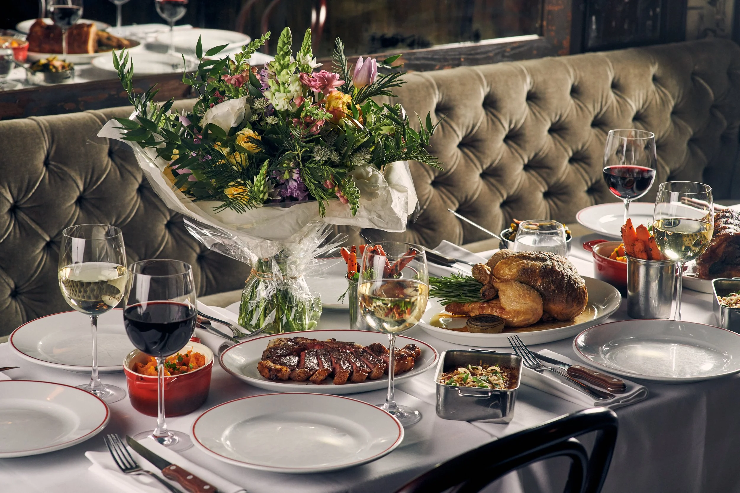 A festive dining table set for a celebration with a centerpiece of a colorful flower bouquet, plates of assorted food including roasted meats, vegetables, and side dishes, and glasses of wine. The table is covered with a white tablecloth and surrounded by upholstered seating.