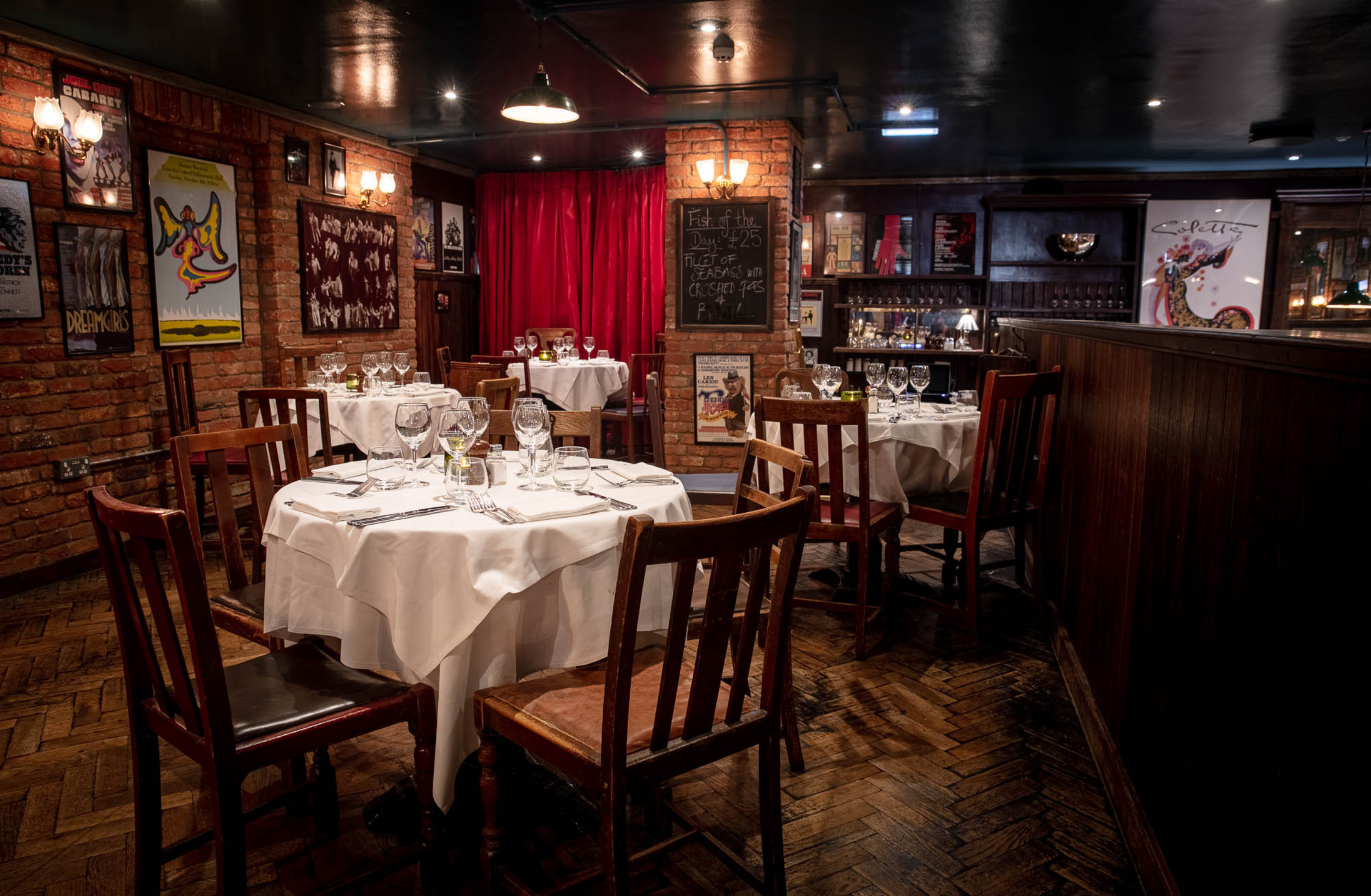 Elegant restaurant dining area with round tables covered in white tablecloths, set with multiple wine glasses, silverware, and napkins, surrounded by wooden chairs, brick walls decorated with posters, a chalkboard with a special menu, and red curtains in the background.