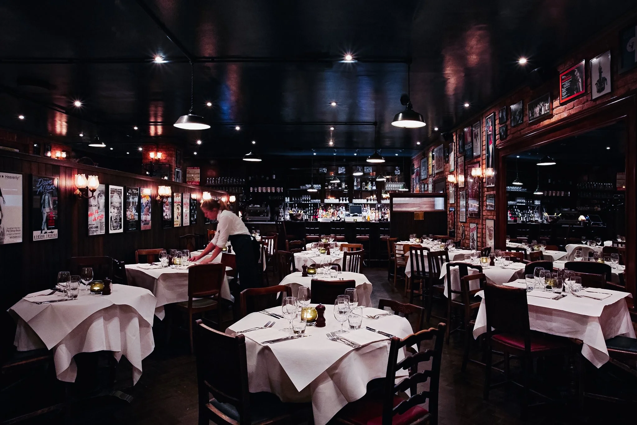 Interior of a dimly lit upscale restaurant with white tablecloth-covered tables, wooden chairs, and framed posters on dark wood-paneled walls. A waitress is setting a table.