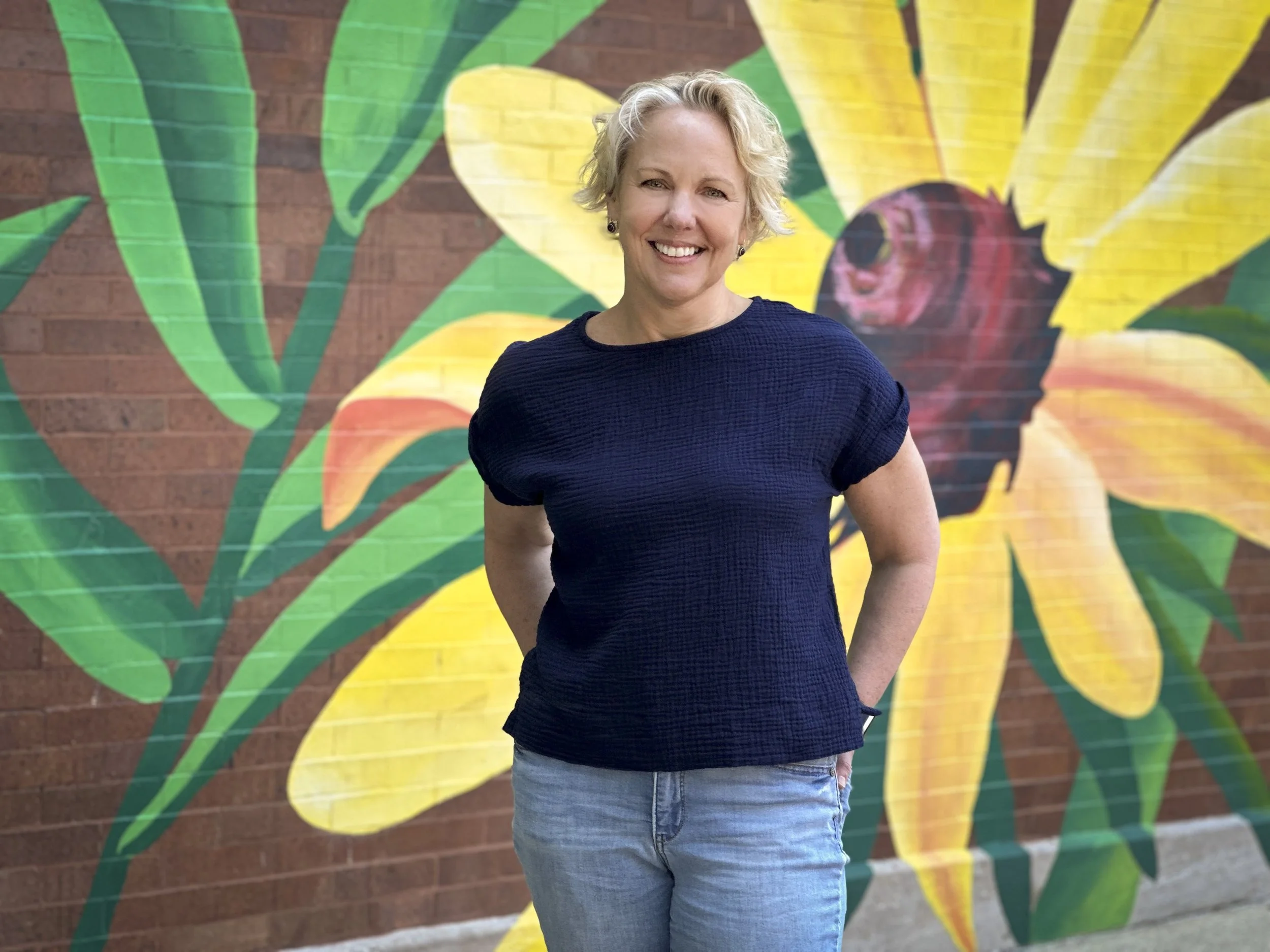 Christine, founder of Stardust Oasis Astrology, standing in front of a mural of a sunflower. Woman with short blond hair wearing a black t-shirt and jeans.