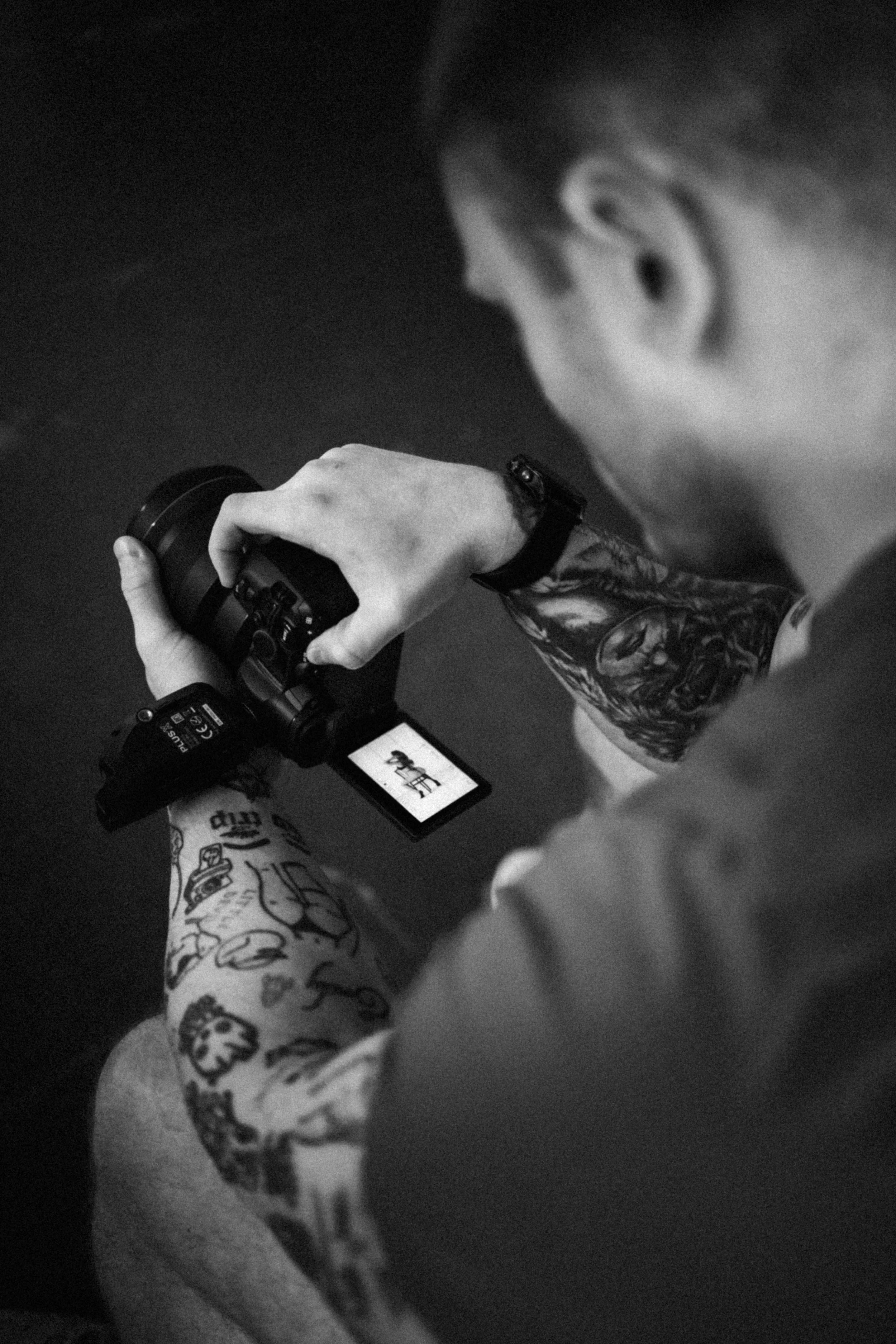 A person with tattoos on their arms is adjusting a camera, looking down at its screen, with a dark background.