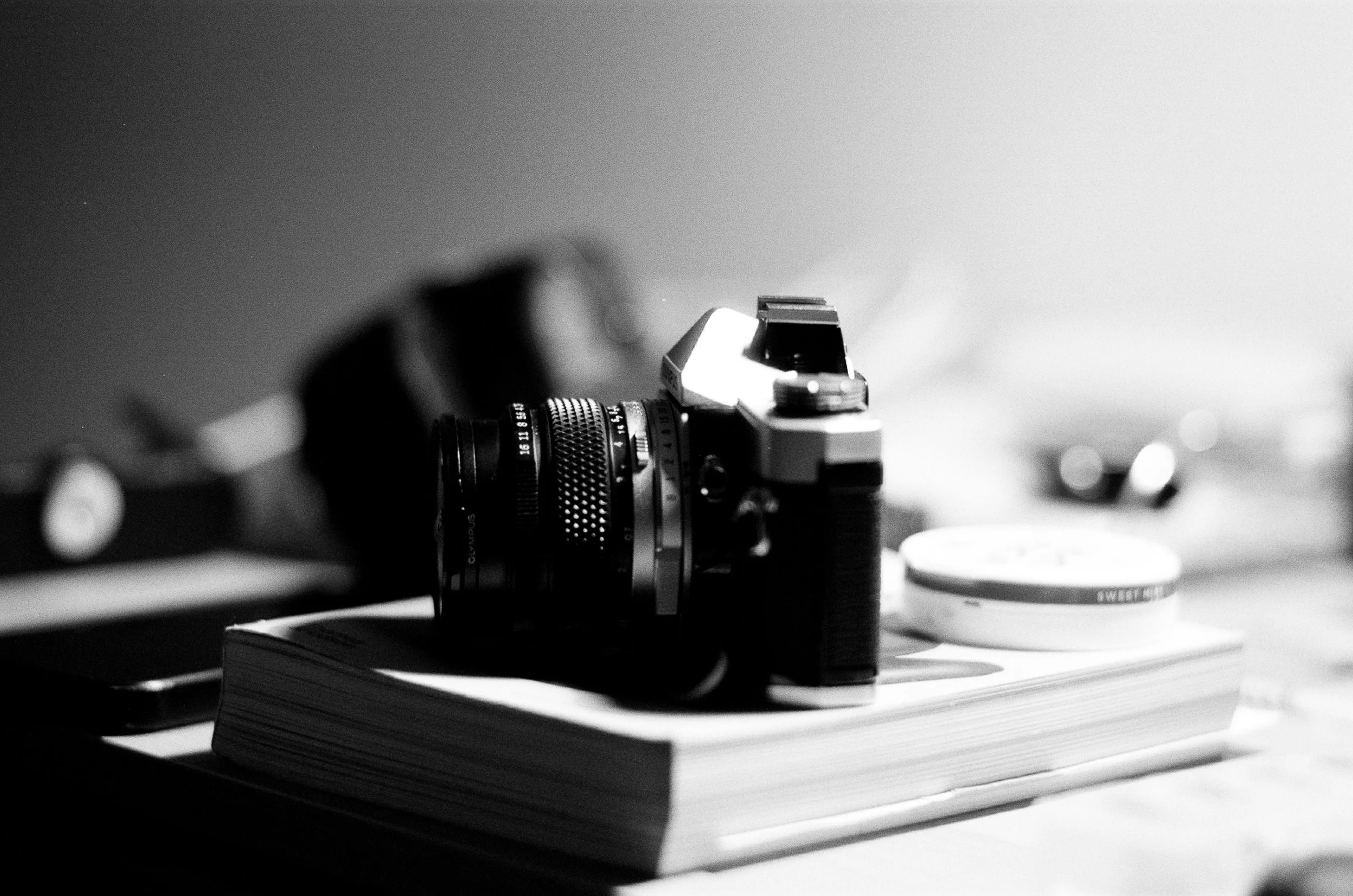 Black and white photo of a camera placed on top of a notebook, with a few other objects blurred in the background.