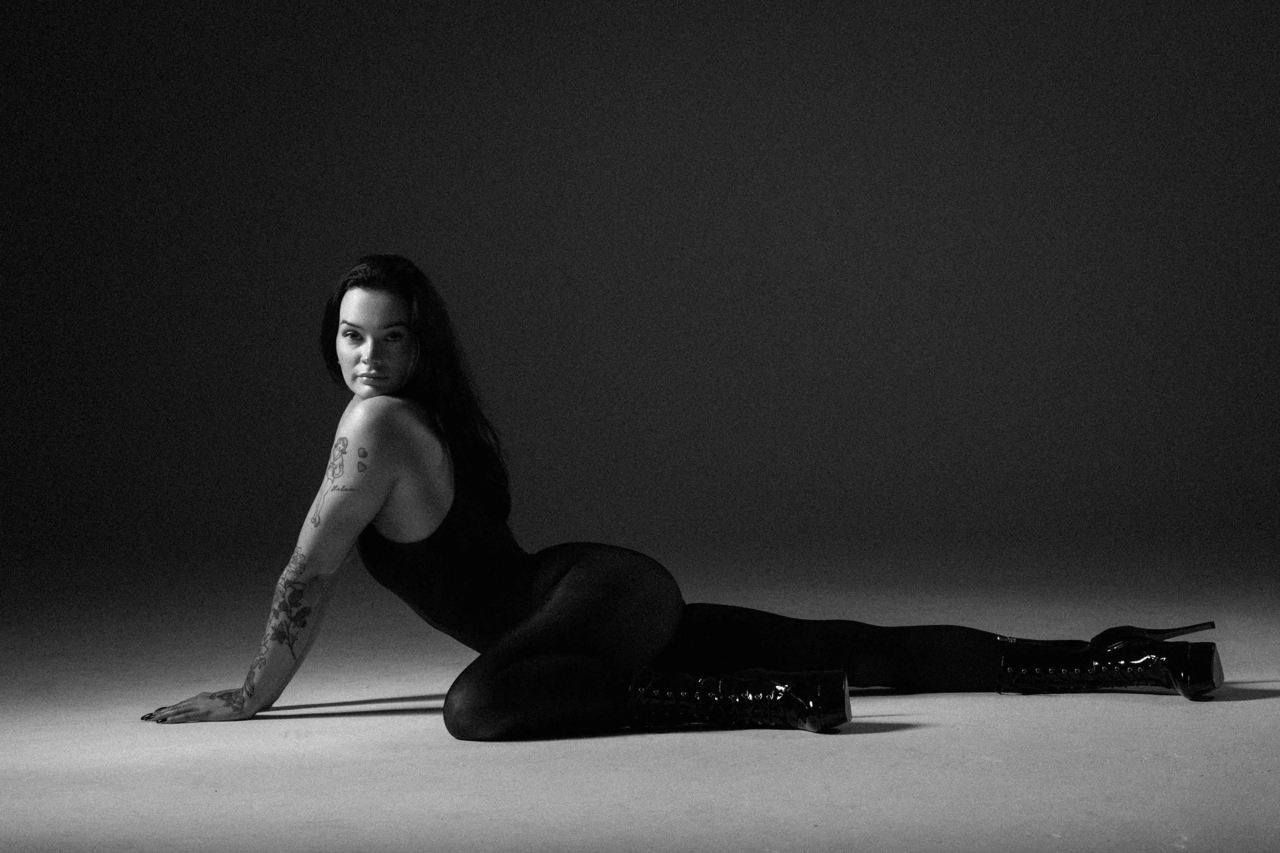 A woman with long dark hair, tattoos on her arm, and light skin, posed on her hands and knees on a flat surface, wearing dark clothing and shiny high-heeled boots, in a black-and-white studio shot.